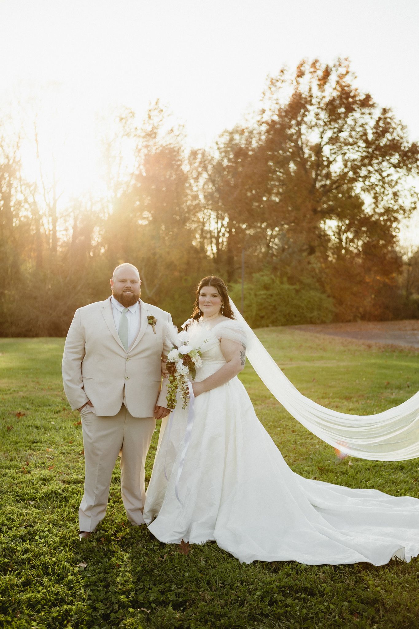 A romantic wedding couple poses in a grassy field during golden hour sunset with a flowing white veil and dress.