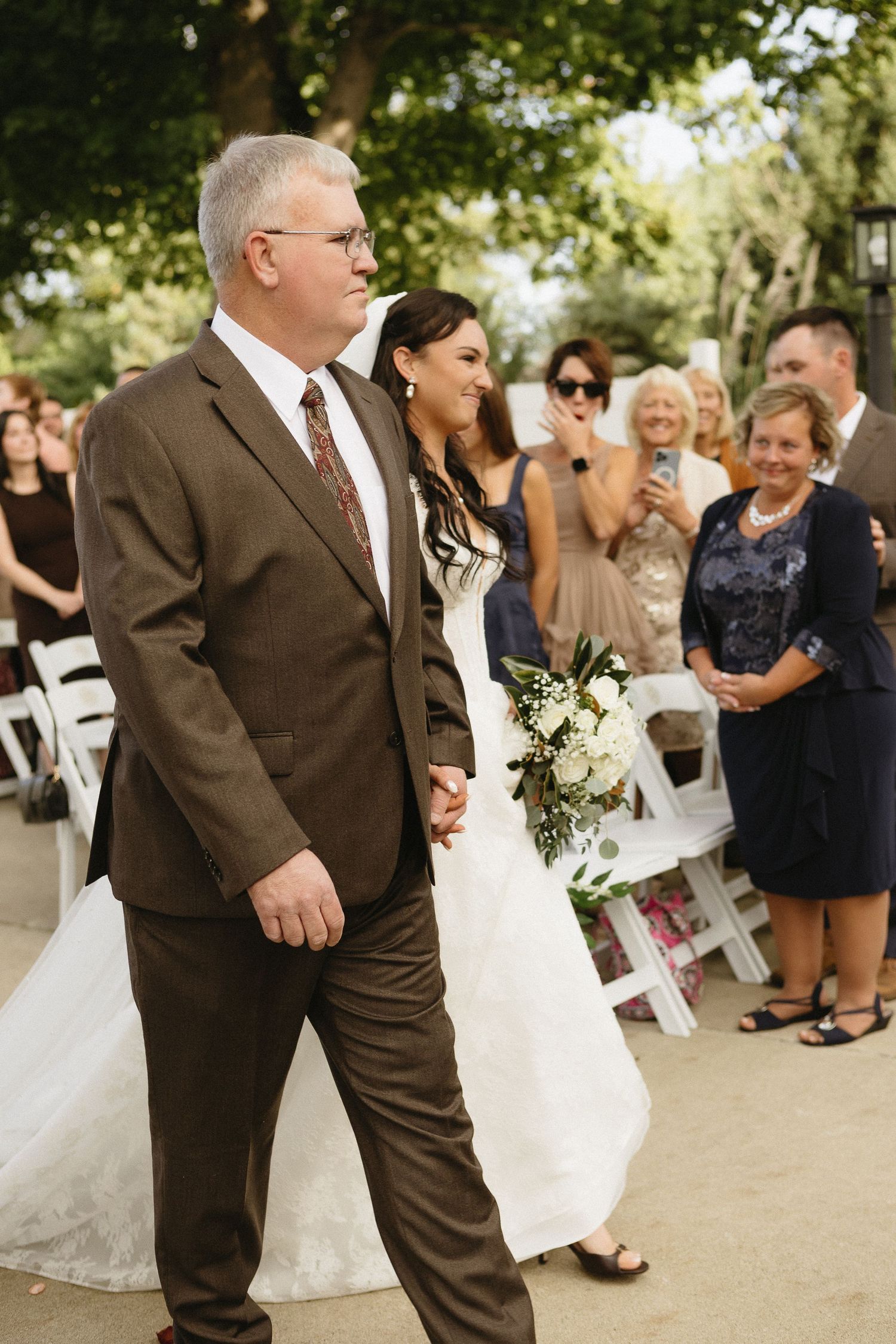Outdoor wedding ceremony with guests seated in white chairs and brown-suited attendants.