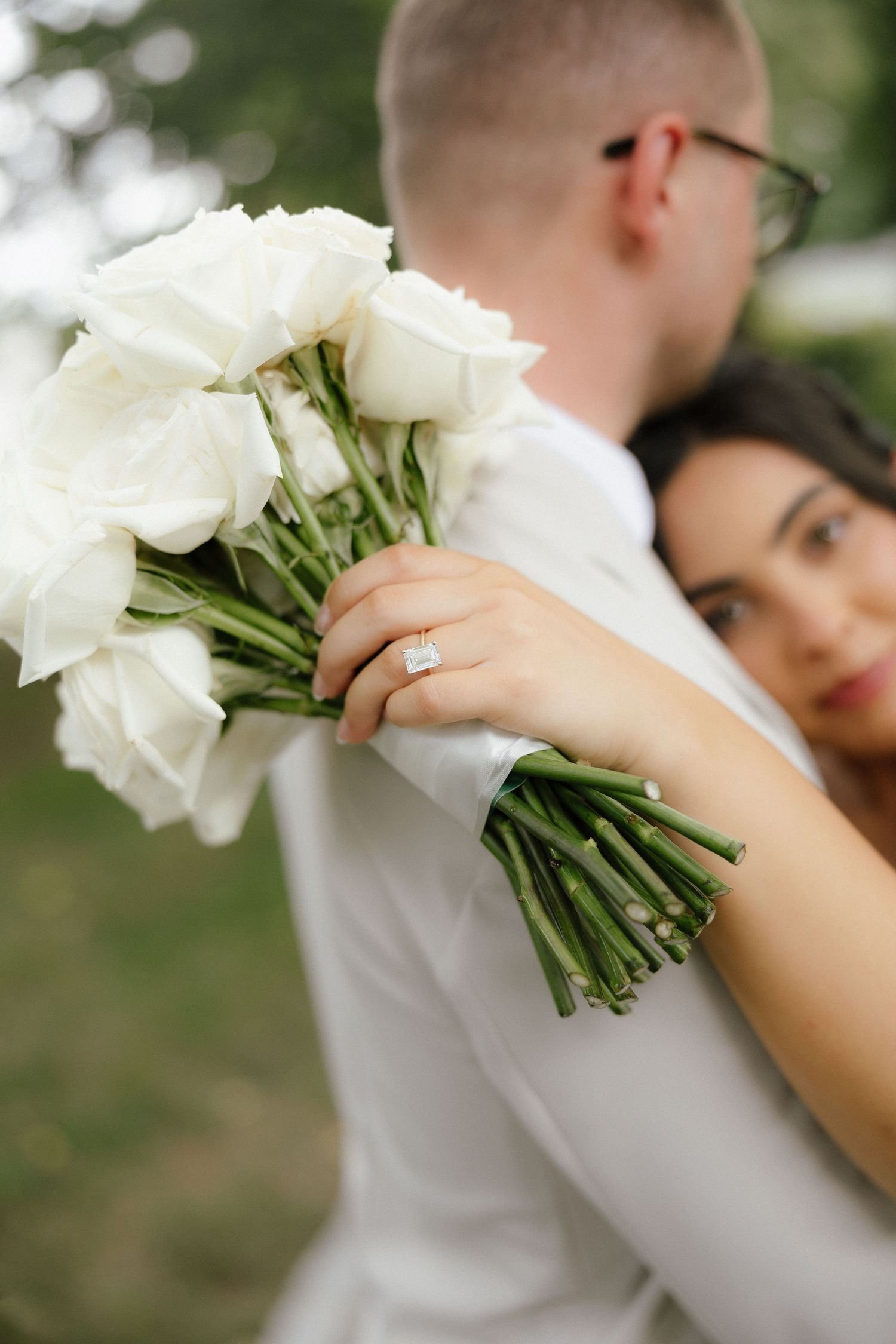 Bride hugs groom while displaying diamond engagement ring and white rose bouquet at outdoor wedding.