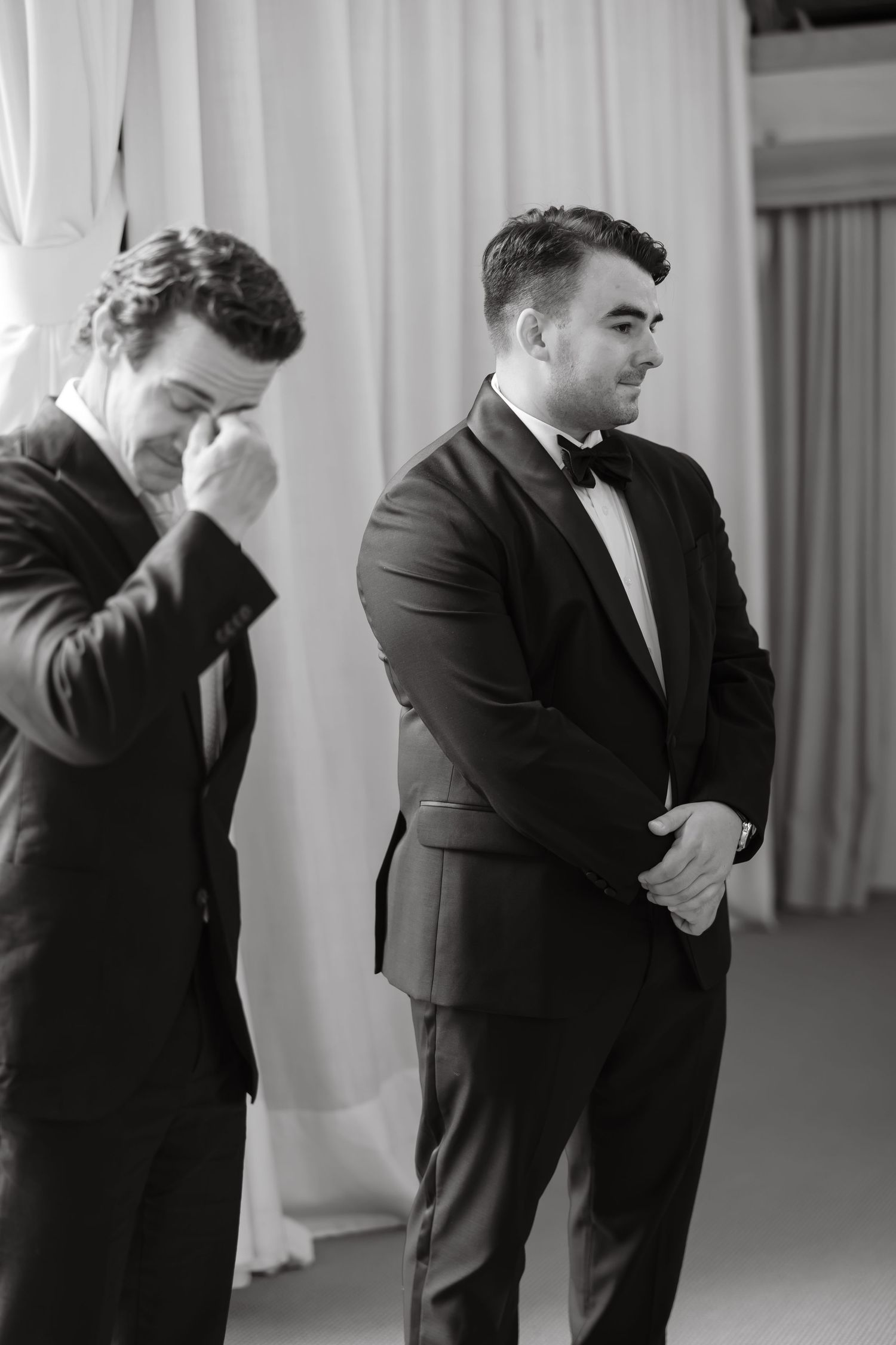 Emotional groom wipes tears while groomsman smiles during wedding ceremony in black and white photo.