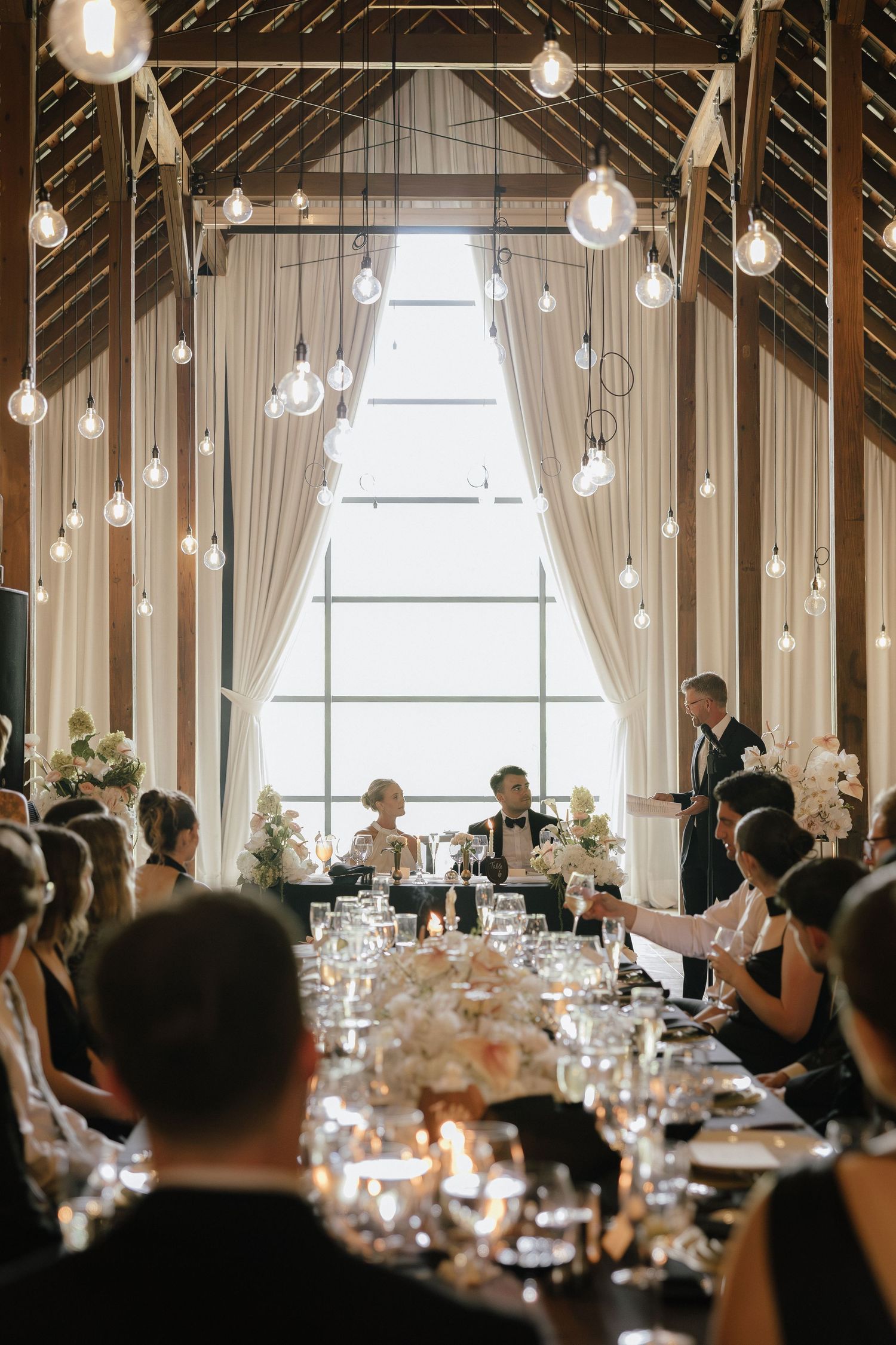 Wedding reception in rustic barn venue with Edison bulb ceiling, long decorated tables and seated guests.