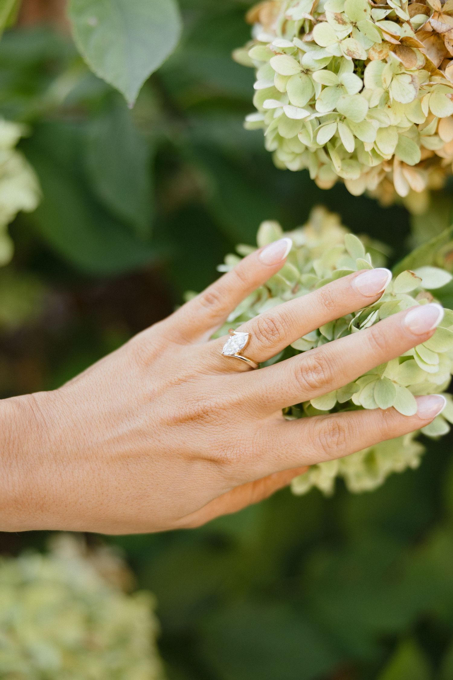 Close-up of engagement ring with emerald-cut diamond on woman's hand near green hydrangea flowers.