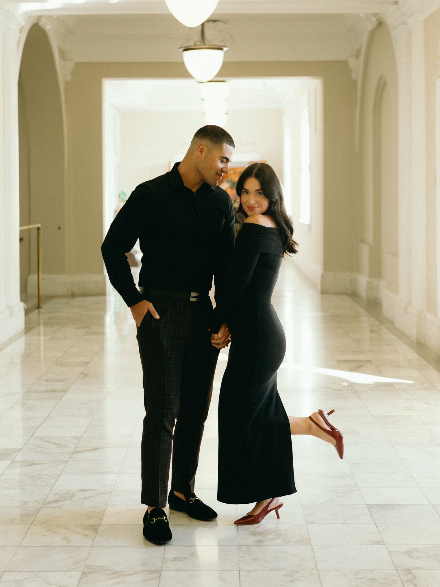A couple in matching black outfits pose elegantly in a bright marble hallway with classic architectural details.