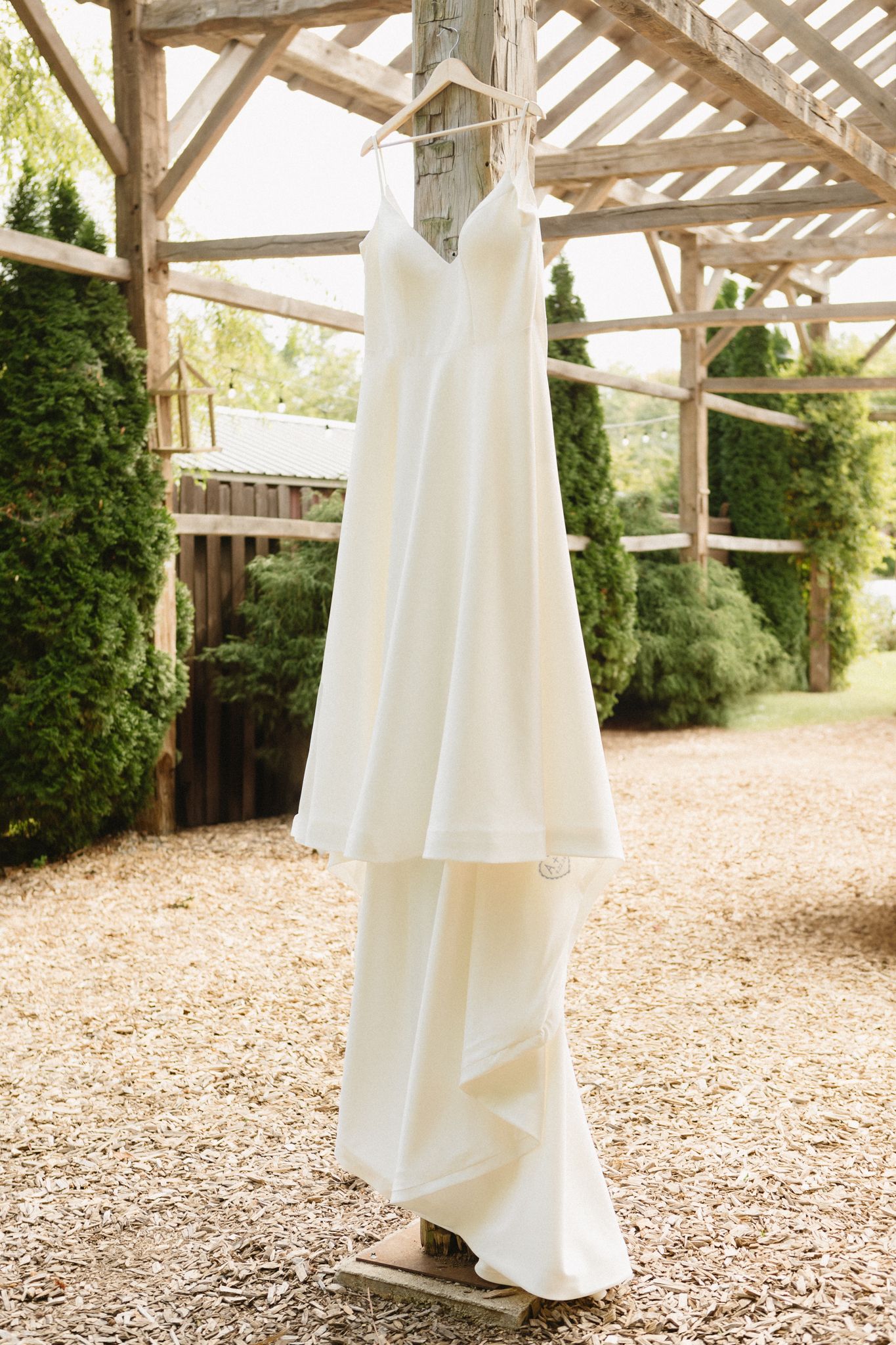 A simple white wedding dress hangs elegantly from a rustic wooden pergola structure in an outdoor garden setting.