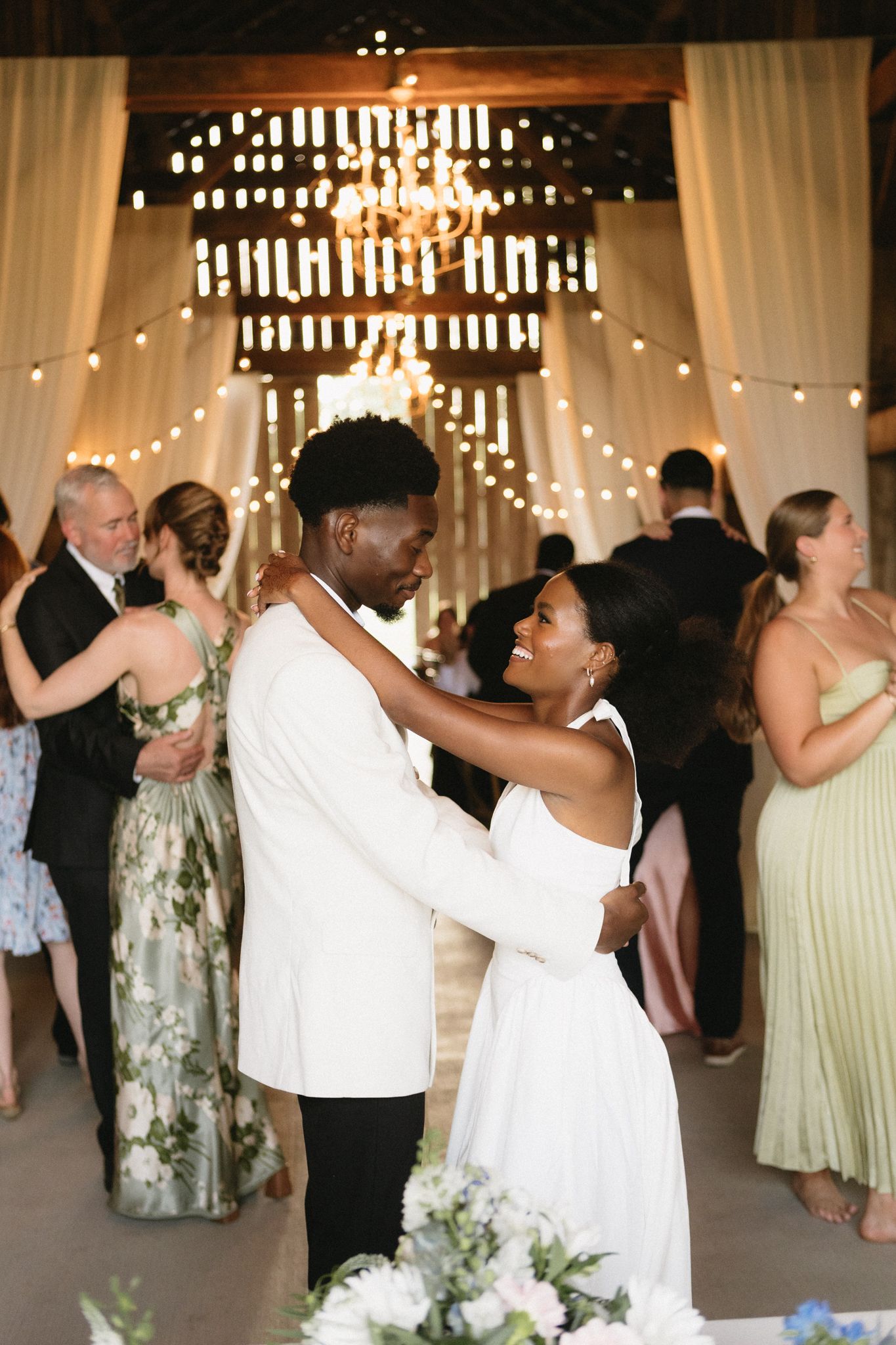 Wedding reception dance party in a rustic barn with string lights and white draping.