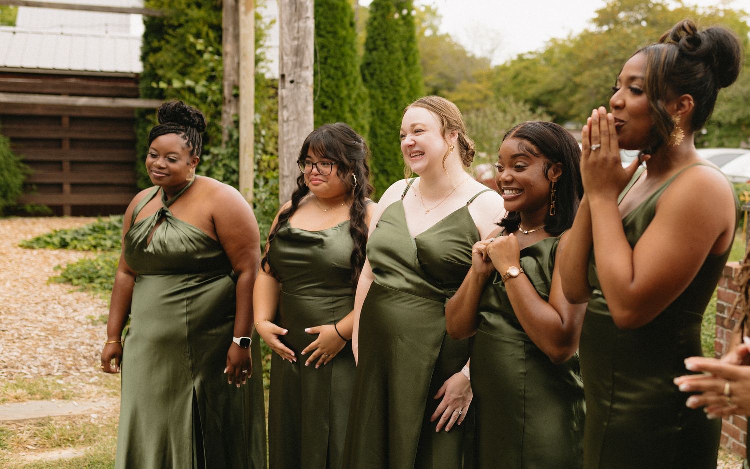 Five bridesmaids in matching olive green dresses share an emotional moment during a wedding celebration outdoors.