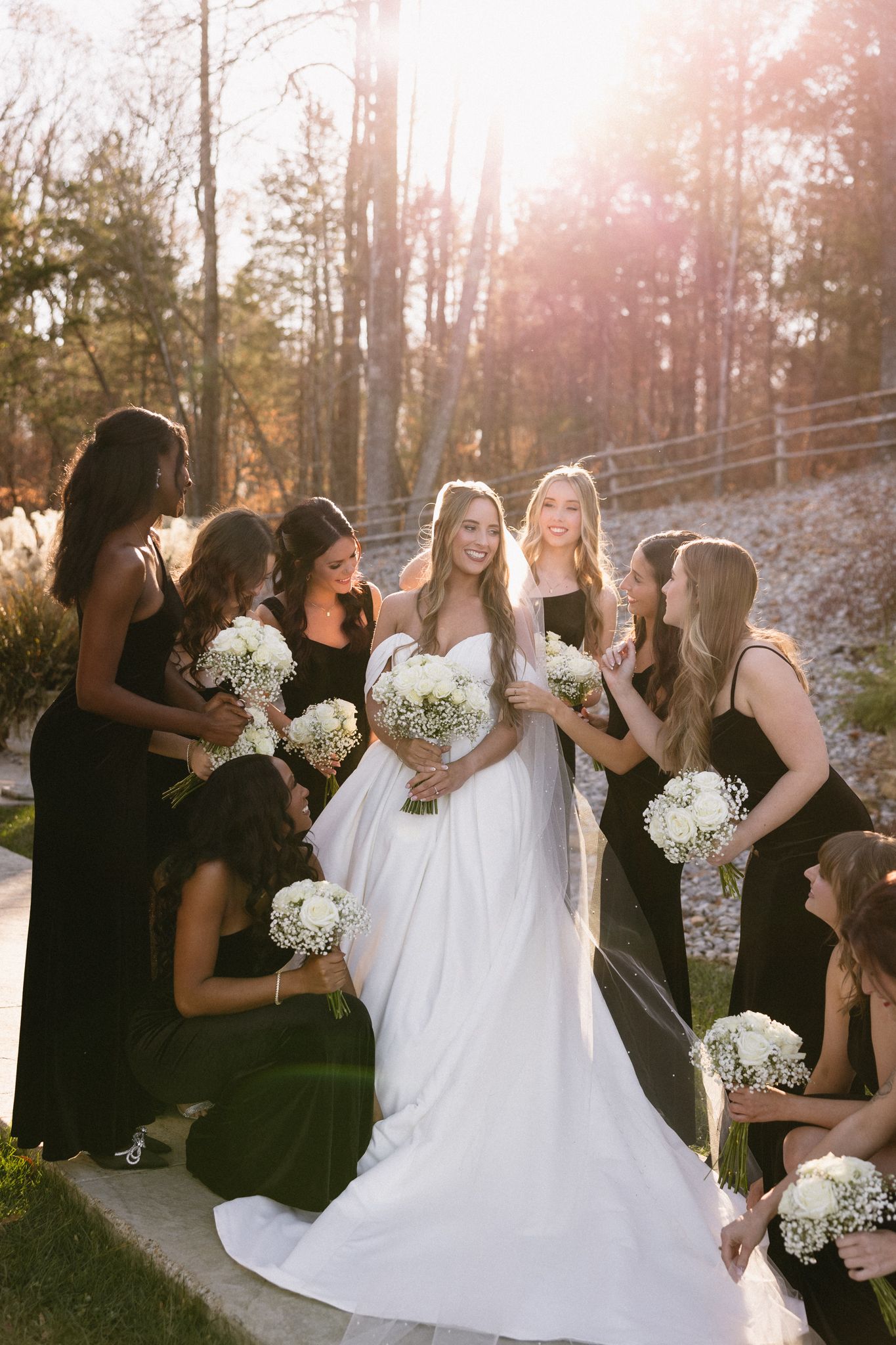 Bridal party in black dresses and white gown pose outdoors in sunlit wooded setting with baby's breath bouquets.
