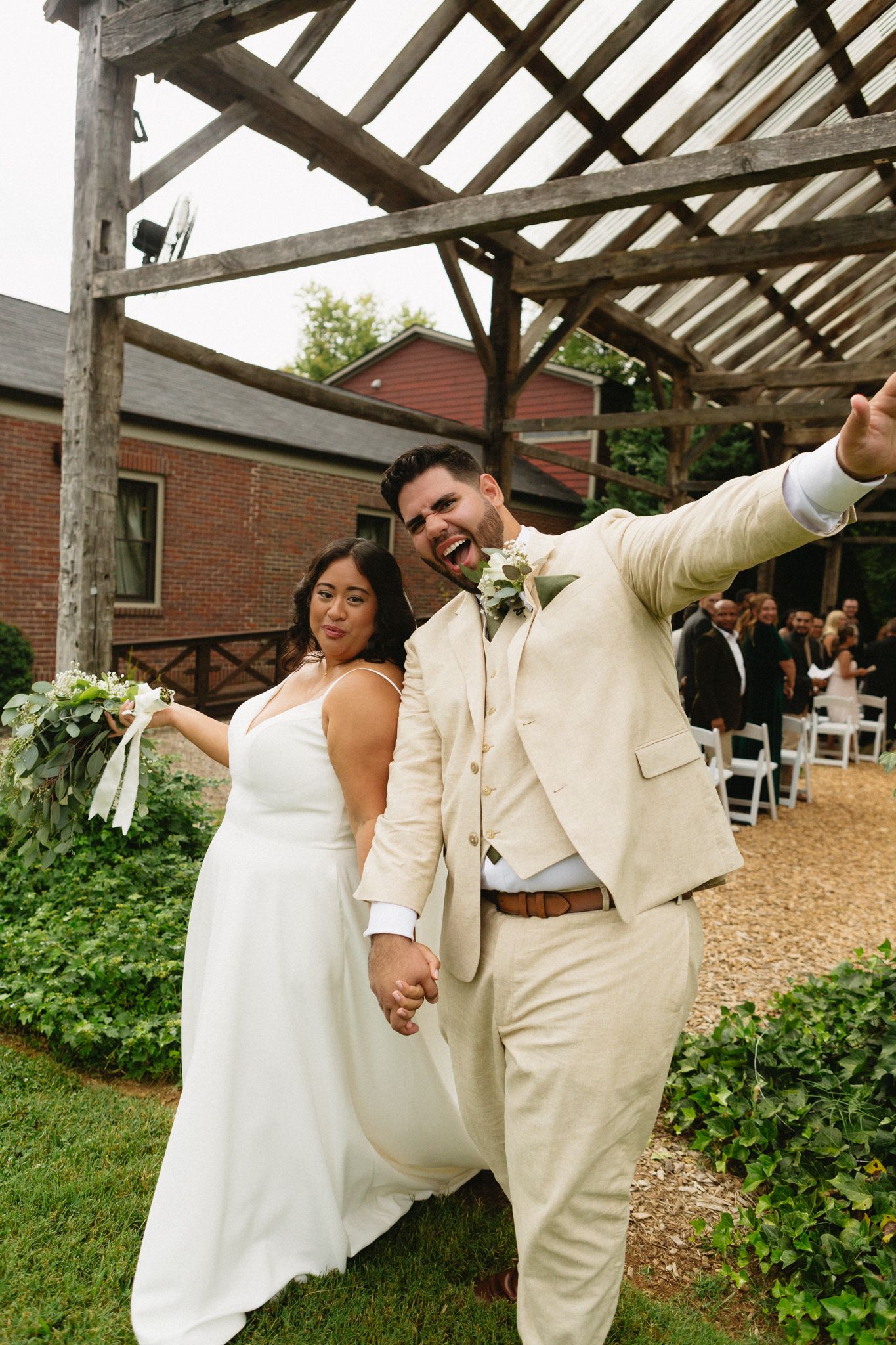 A joyful wedding couple strikes a playful pose under a rustic covered outdoor venue with exposed wooden beams.