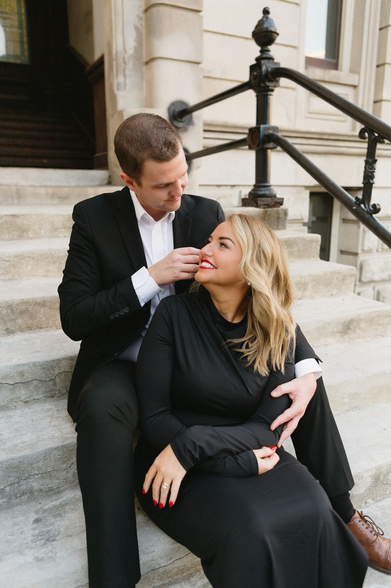 An elegantly dressed couple shares an intimate moment while sitting on outdoor steps of a building.