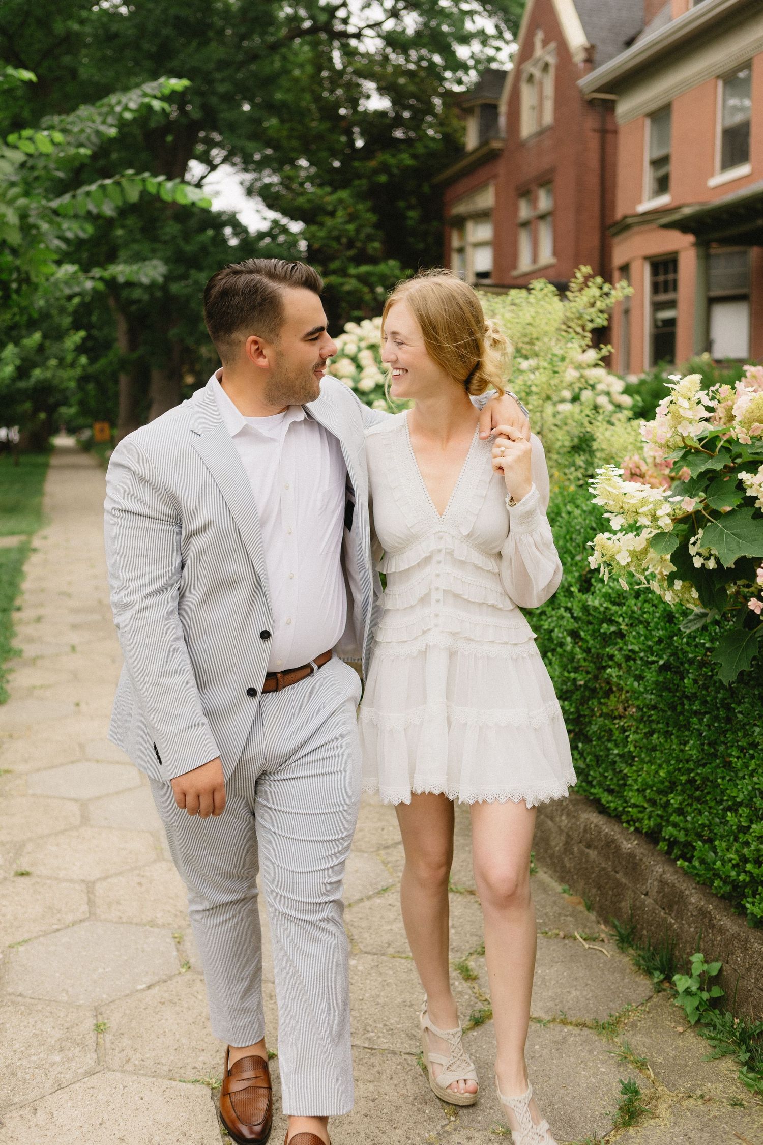 Couple in elegant white dress and seersucker suit walking and smiling on a garden pathway.