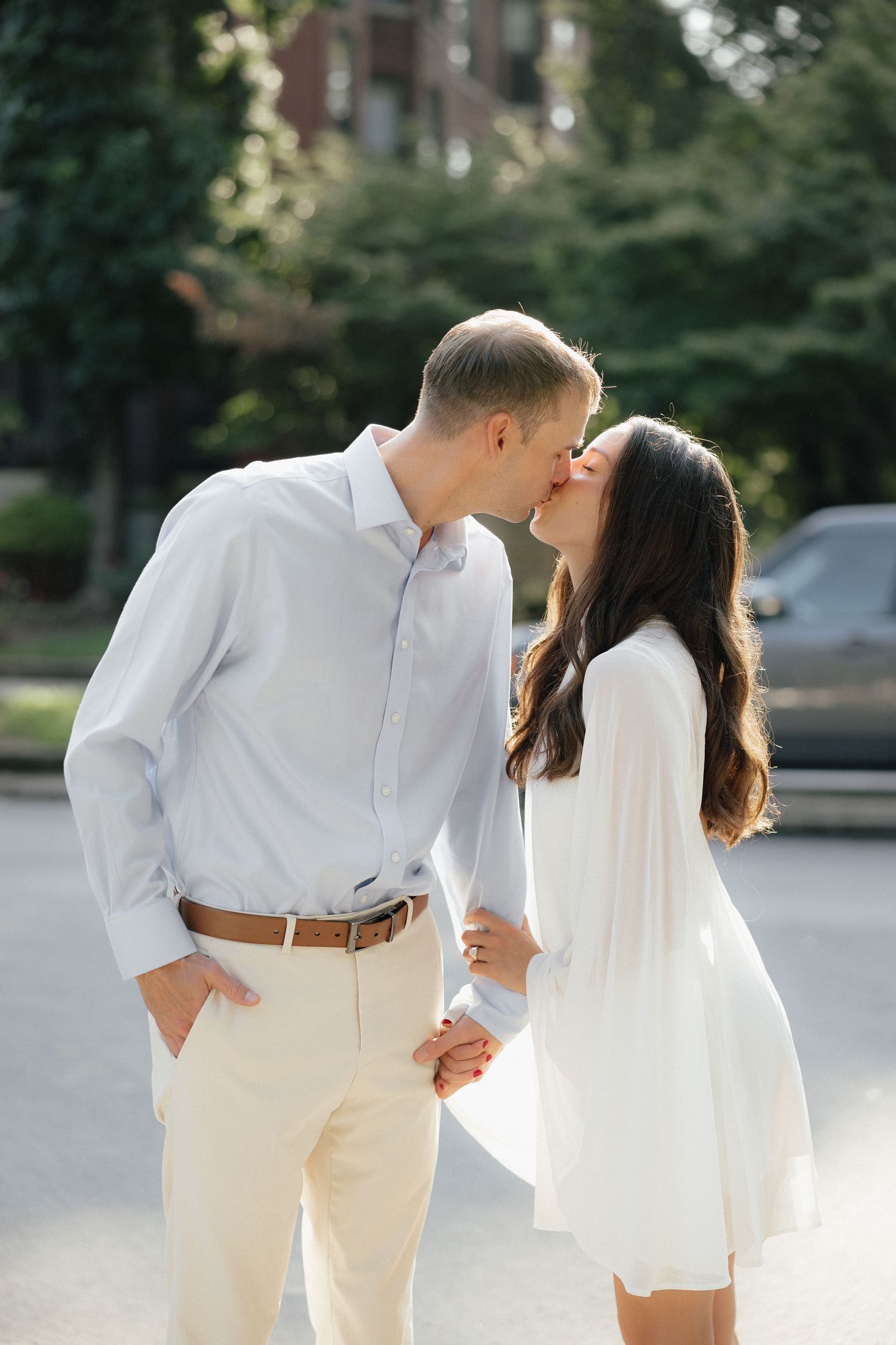 Couple sharing a romantic kiss outdoors, woman in white mini dress and man in light blue shirt holding hands.
