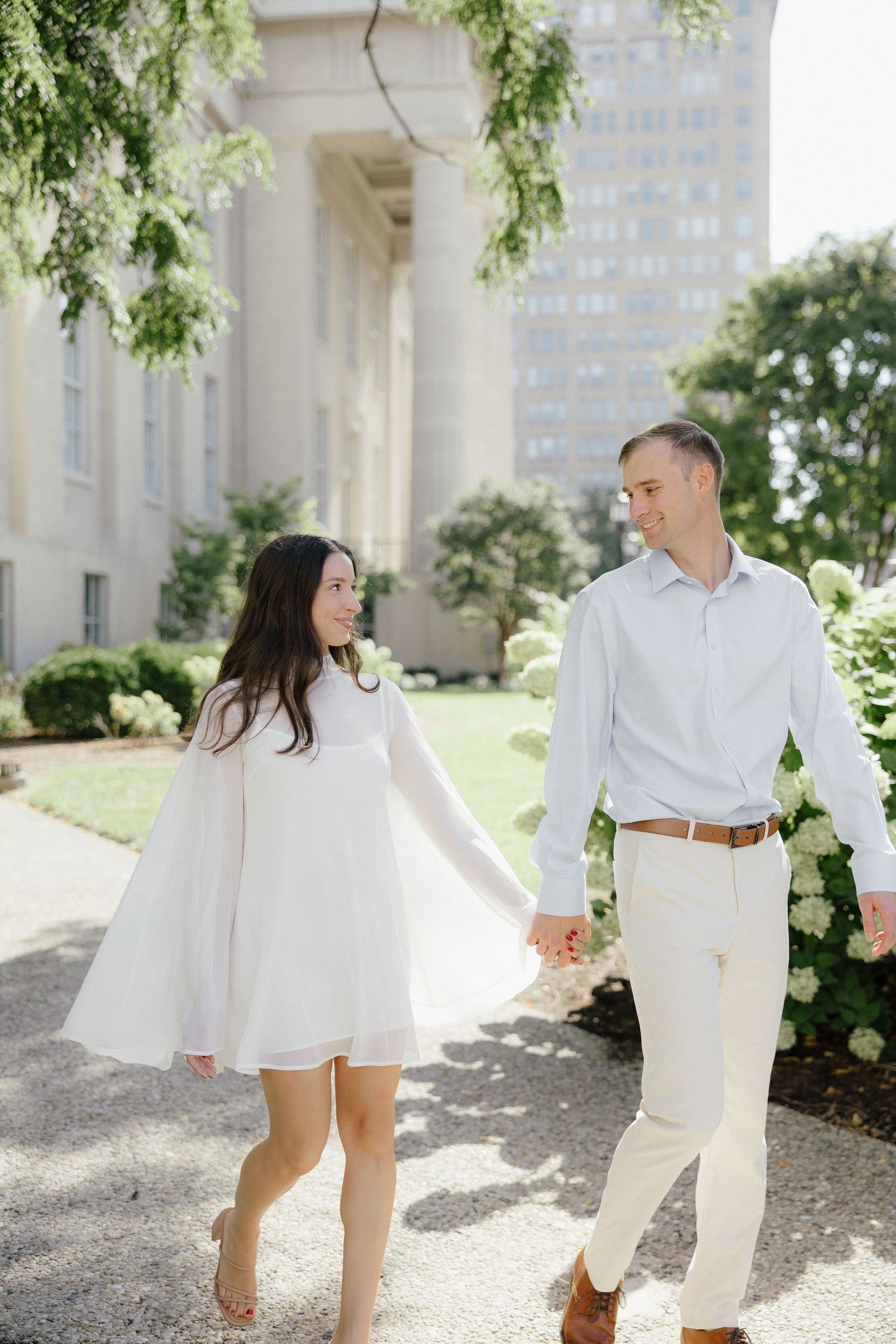 Couple holding hands and walking past white hydrangeas near a classic building with tall columns.