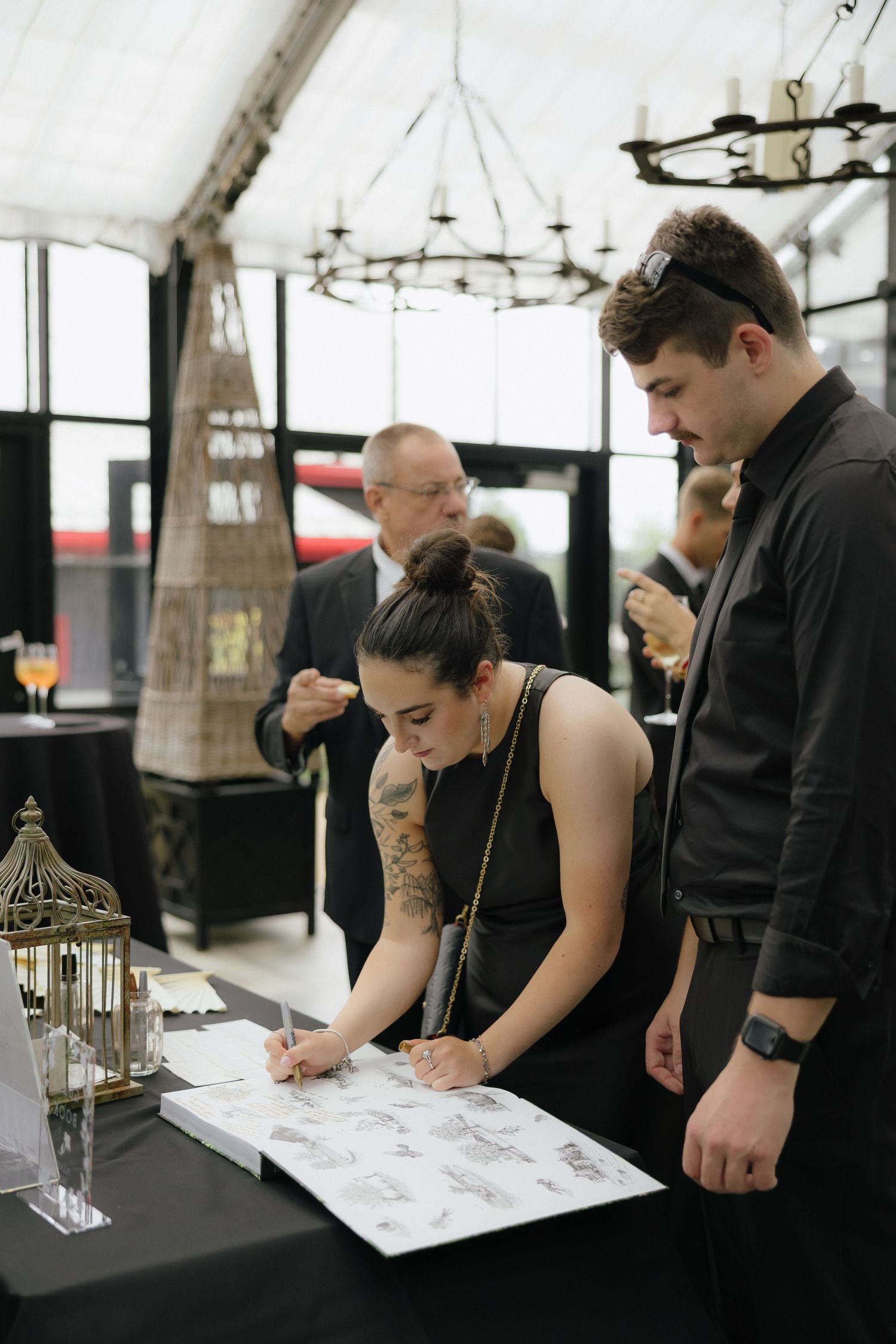 Wedding guest in black dress signs guestbook at elegant reception with chandelier backdrop.