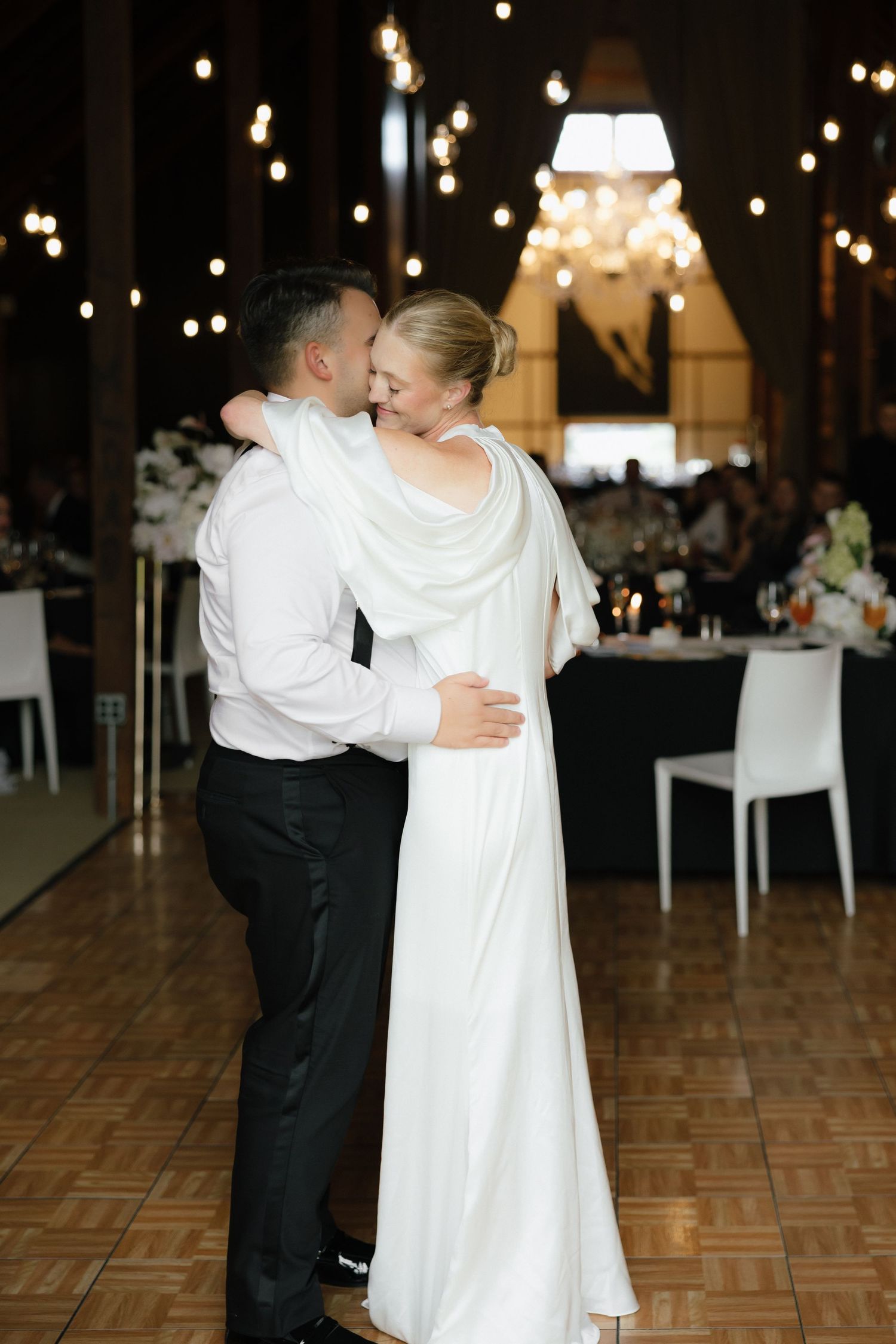 Bride and groom share intimate first dance in warmly lit reception hall with chandelier overhead.