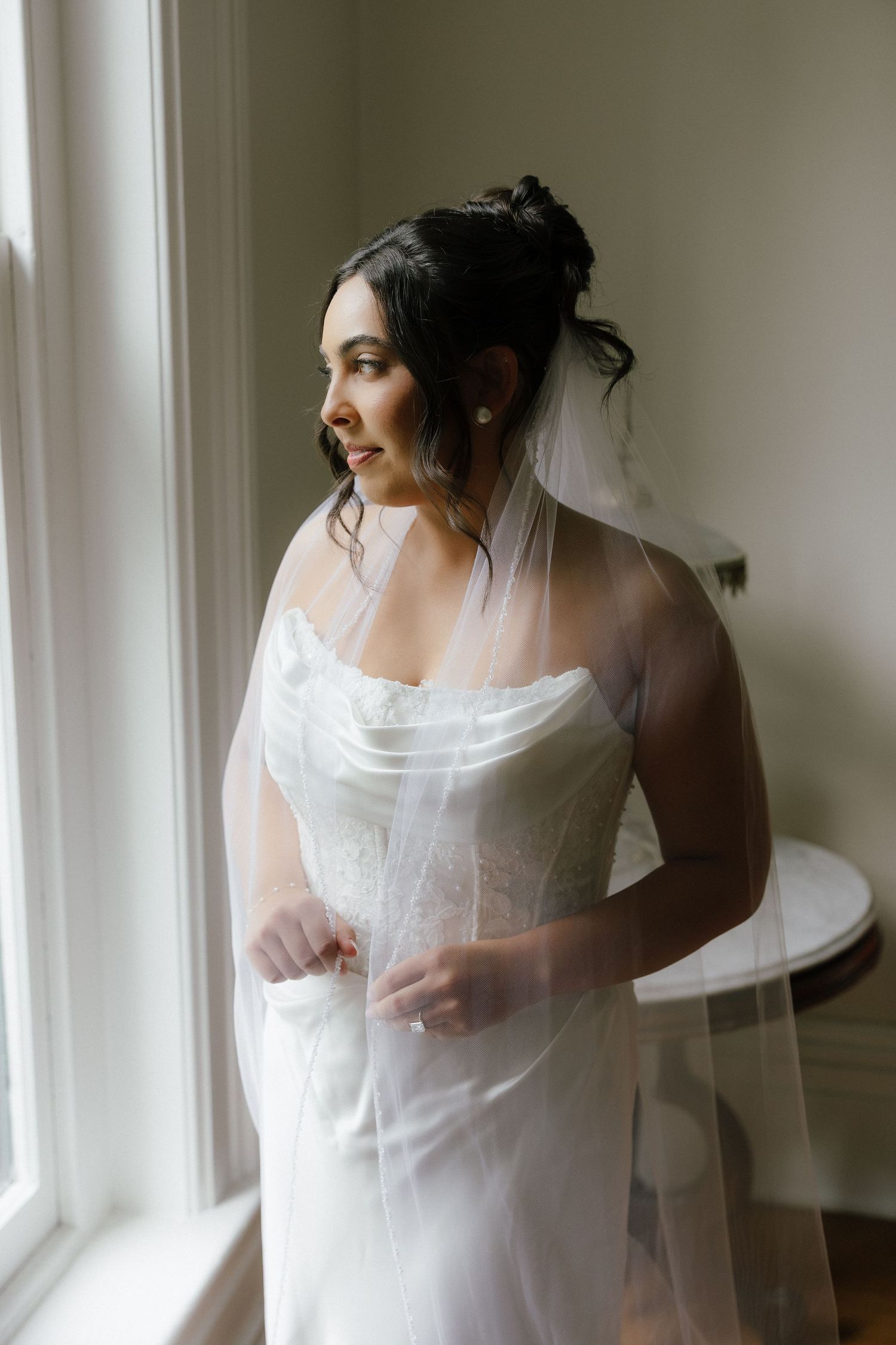 Bride in white lace wedding gown and veil gazes out window, hair styled in elegant updo.