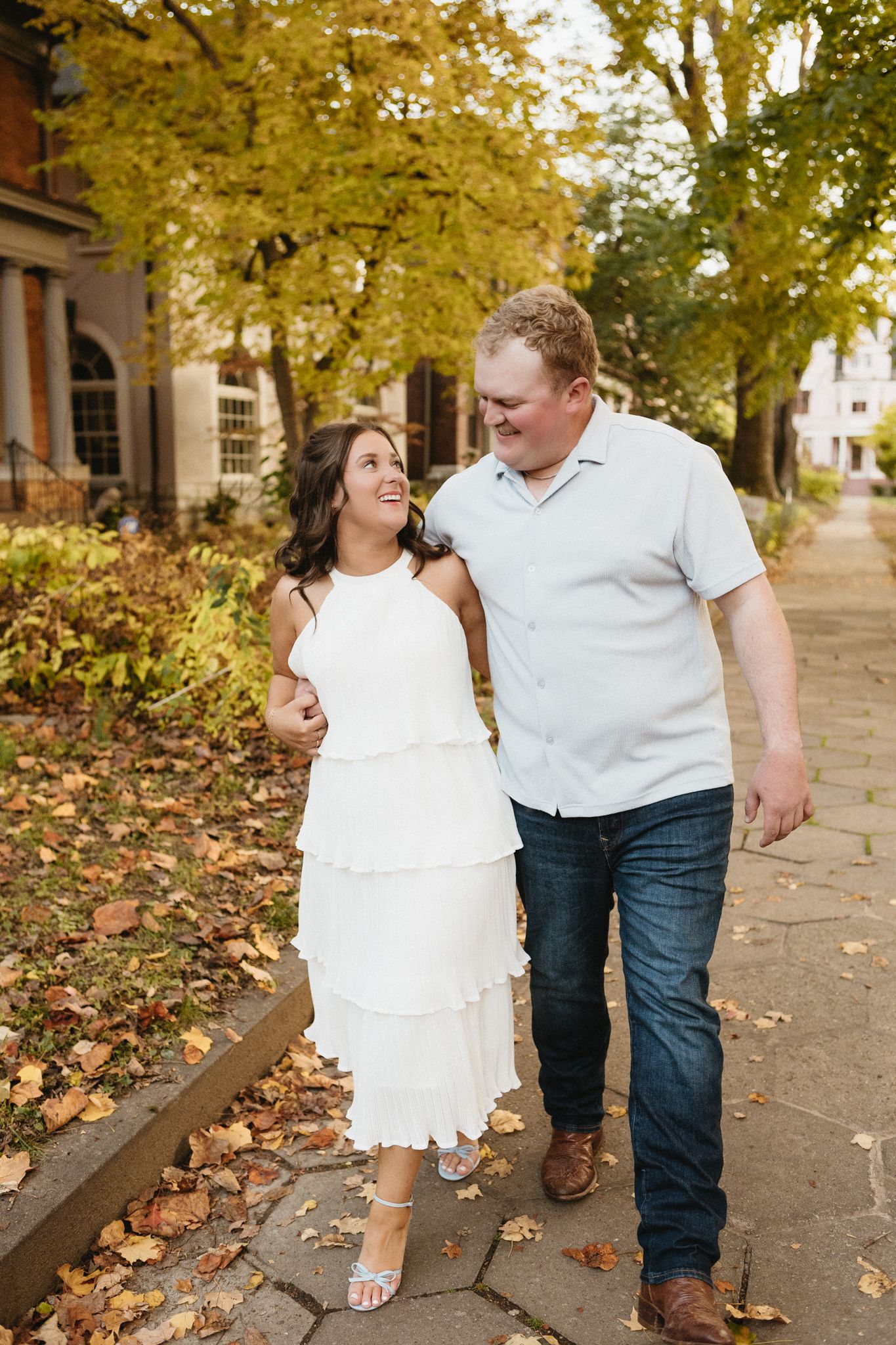 A couple walks together on a leaf-covered sidewalk during fall with golden trees overhead.