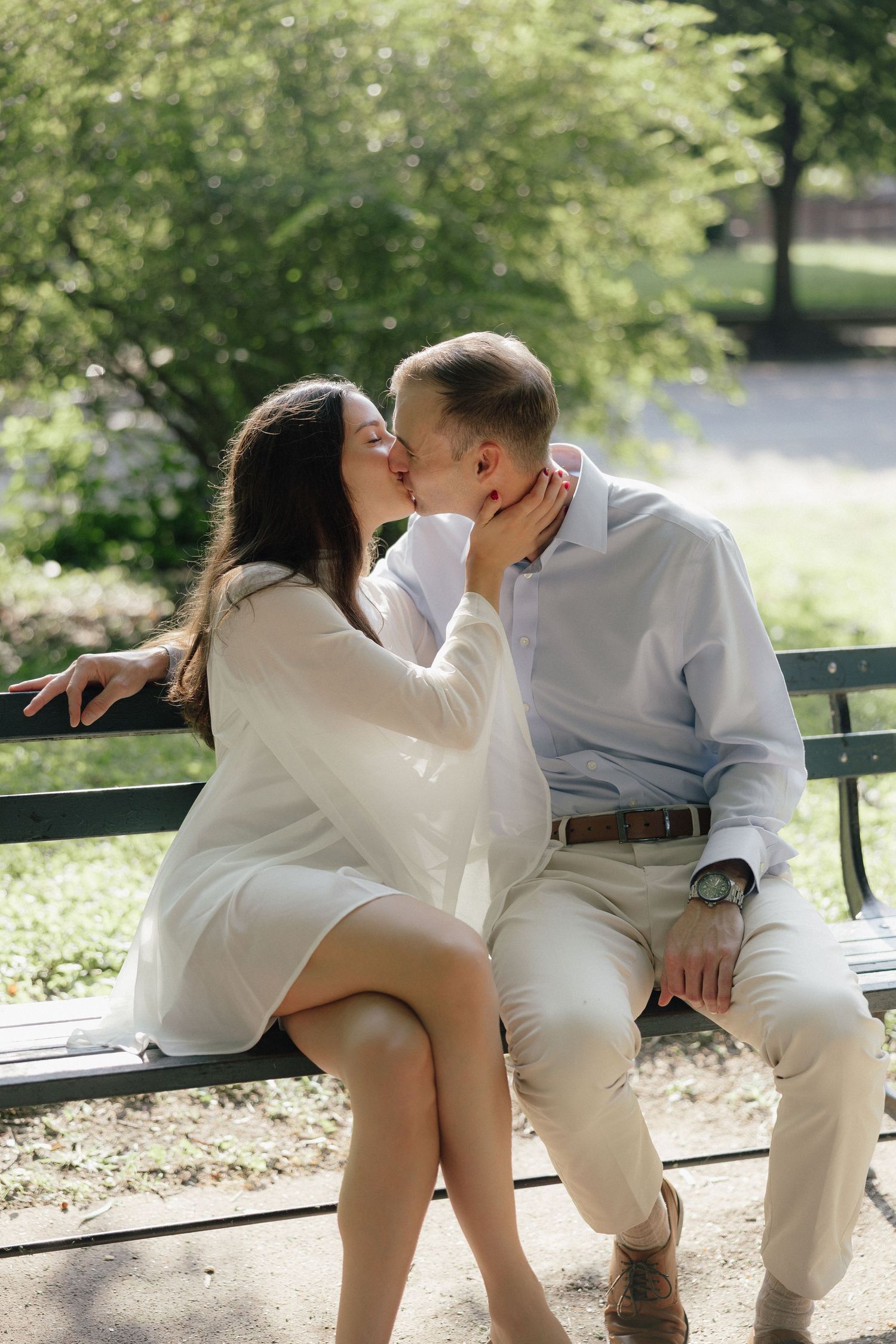 A couple shares a romantic kiss on a park bench surrounded by lush green trees on a sunny day.