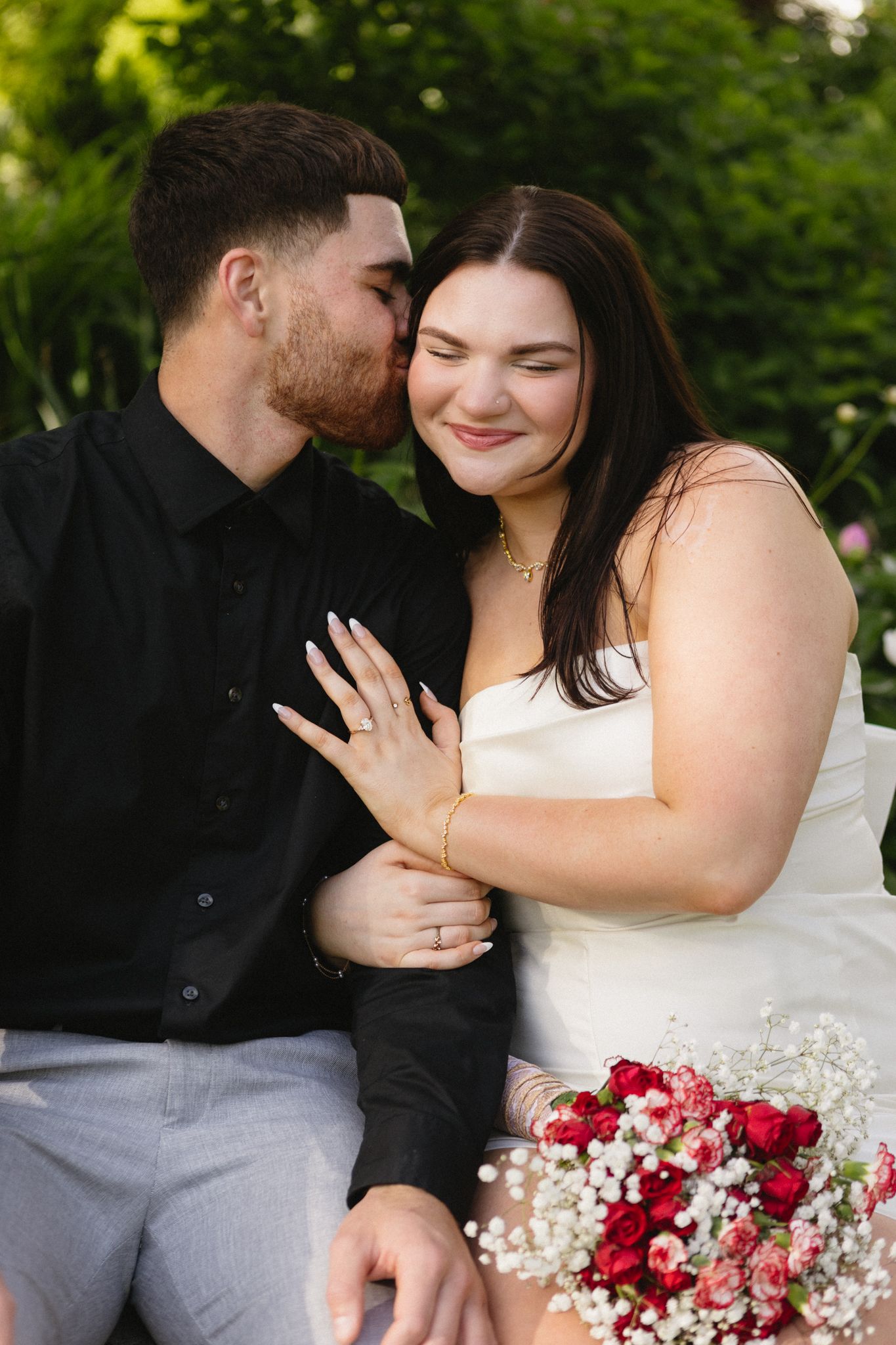 An engaged couple shares tender moments while holding a bouquet of red and pink roses with baby's breath.