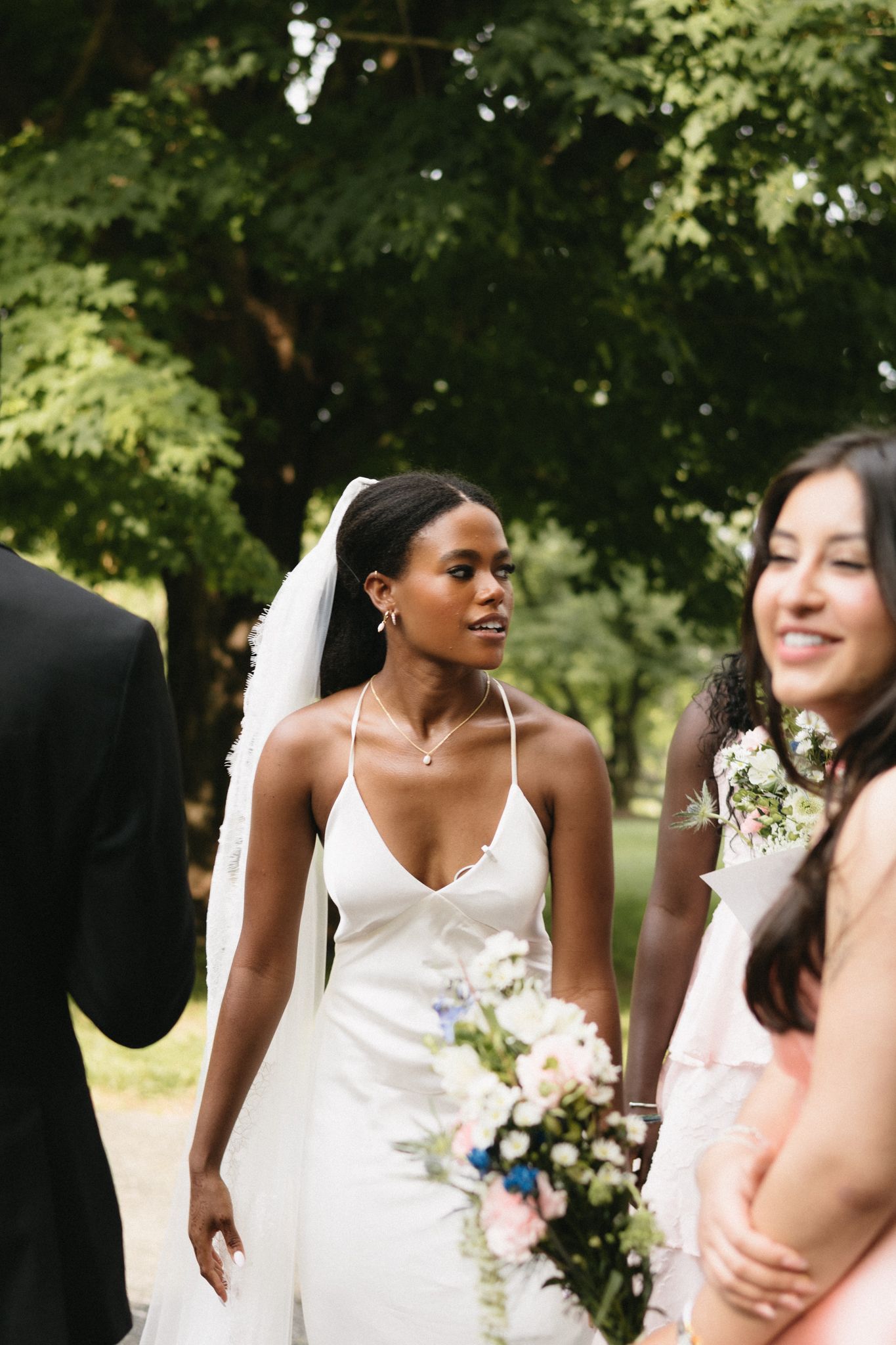 An elegant wedding party gathers outdoors under trees during a summer celebration.
