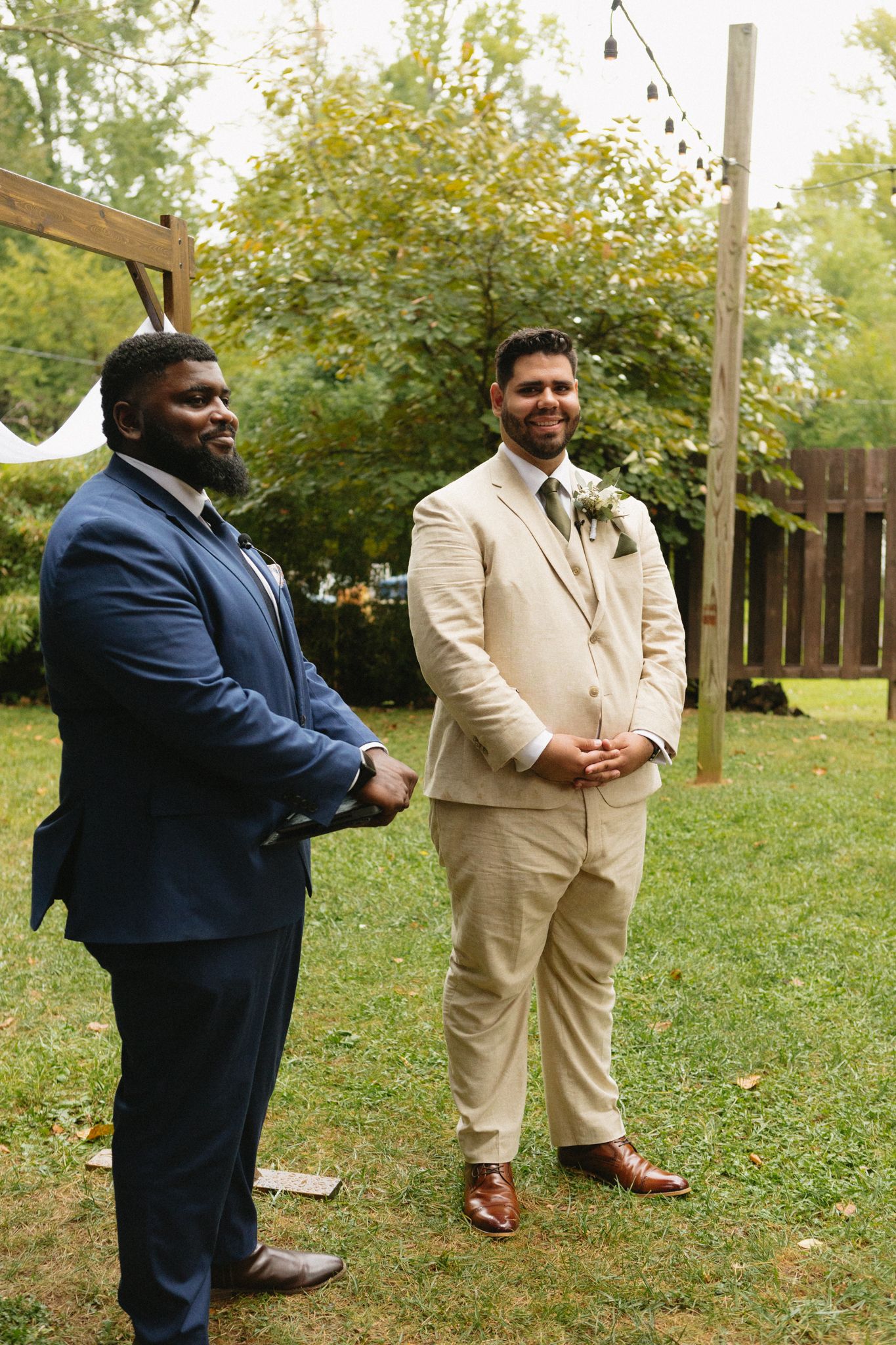 Two men standing in formal attire at an outdoor garden wedding ceremony with string lights overhead.