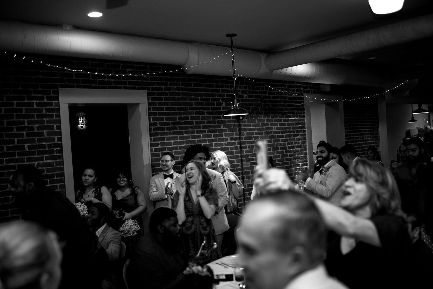 Black and white image of a crowded club venue with performers on stage and audience members in a dimly lit brick room.