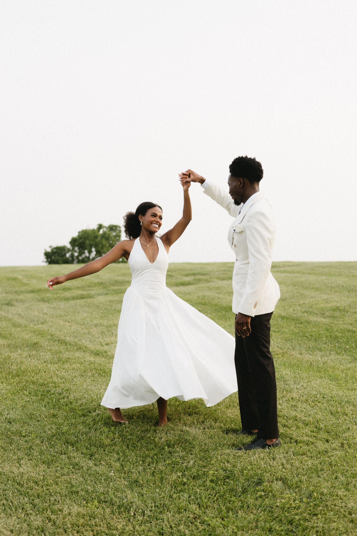 A couple in white formal wear dances joyfully on a green lawn.