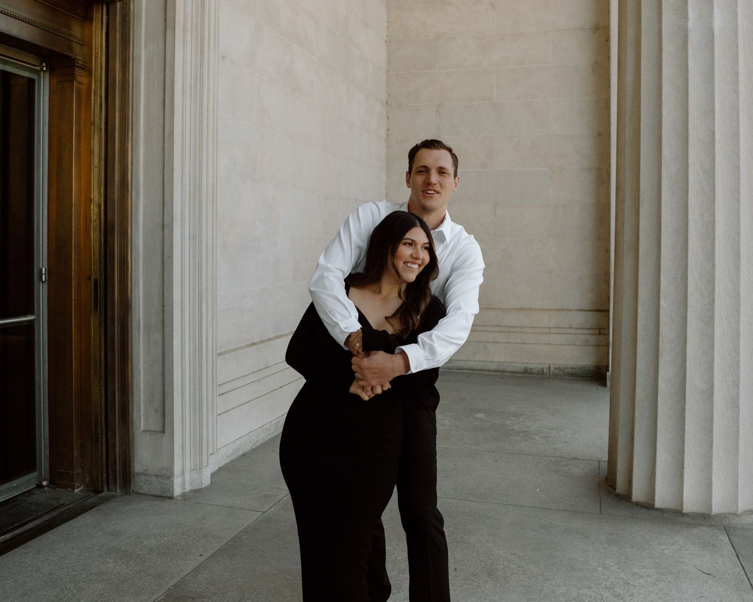 Engaged couple in downtown Louisville, with the woman smiling and showing her engagement ring while embracing her fiancé