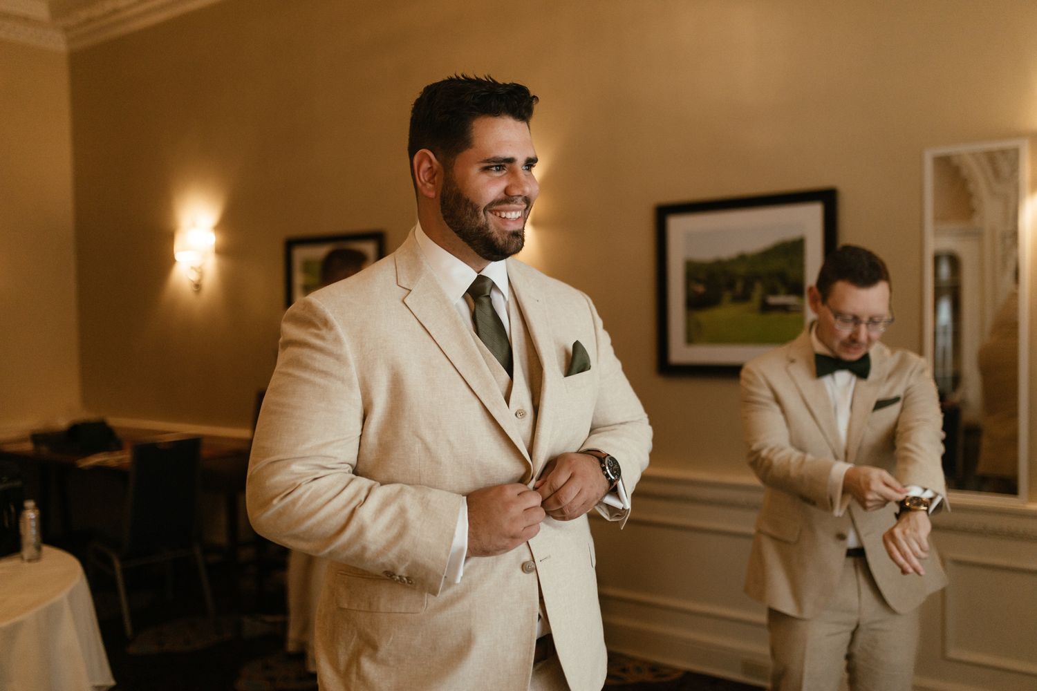 Wedding party members in cream colored suits prepare in an elegant room with warm lighting and framed artwork.