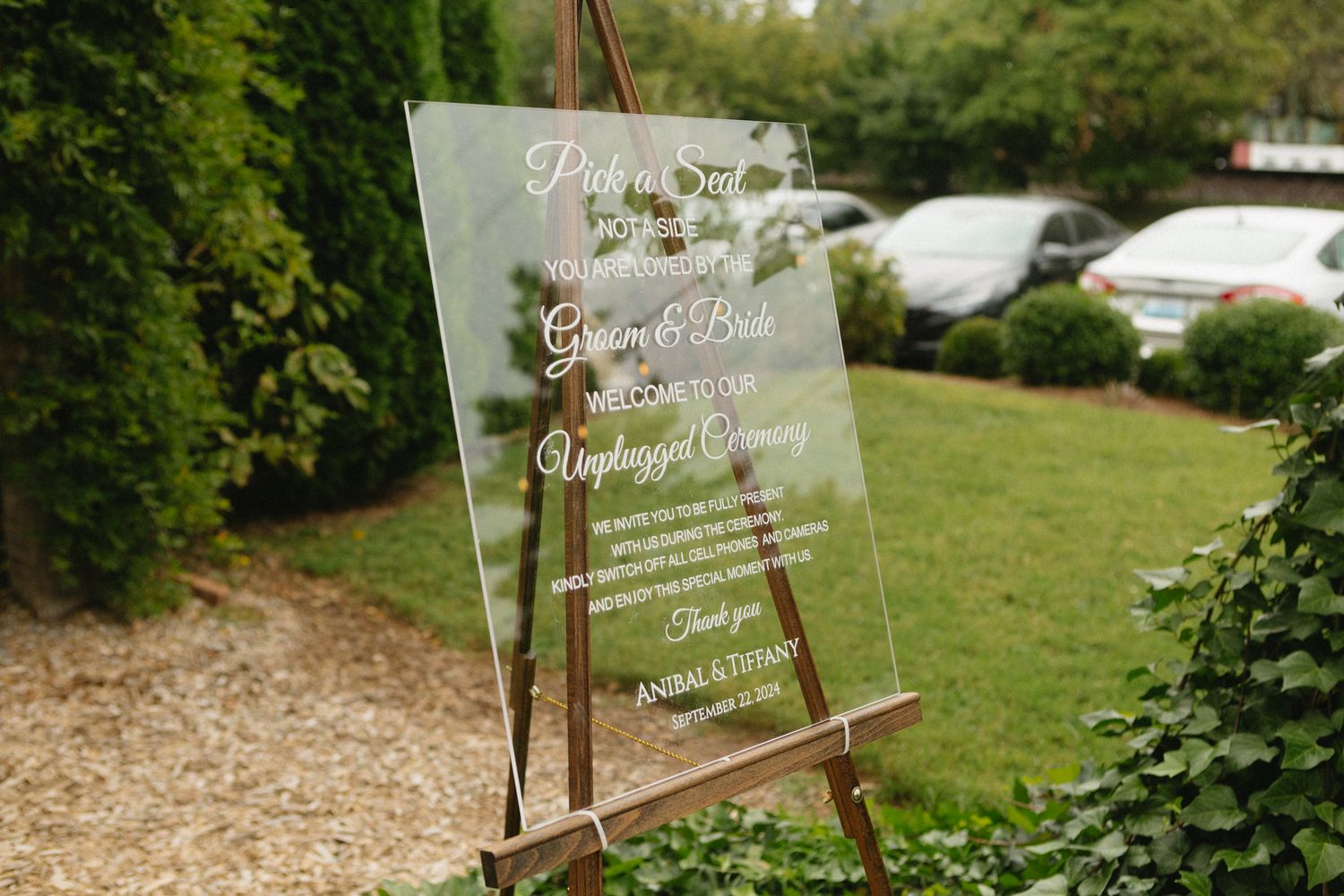Clear acrylic wedding sign displayed on wooden easel in outdoor garden setting with green foliage.