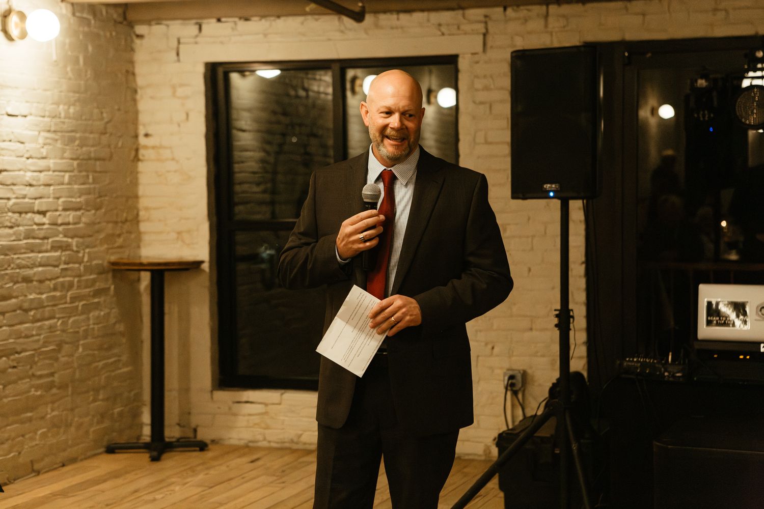 A speaker in a black suit stands in a rustic venue with white brick walls and warm lighting during an evening event.
