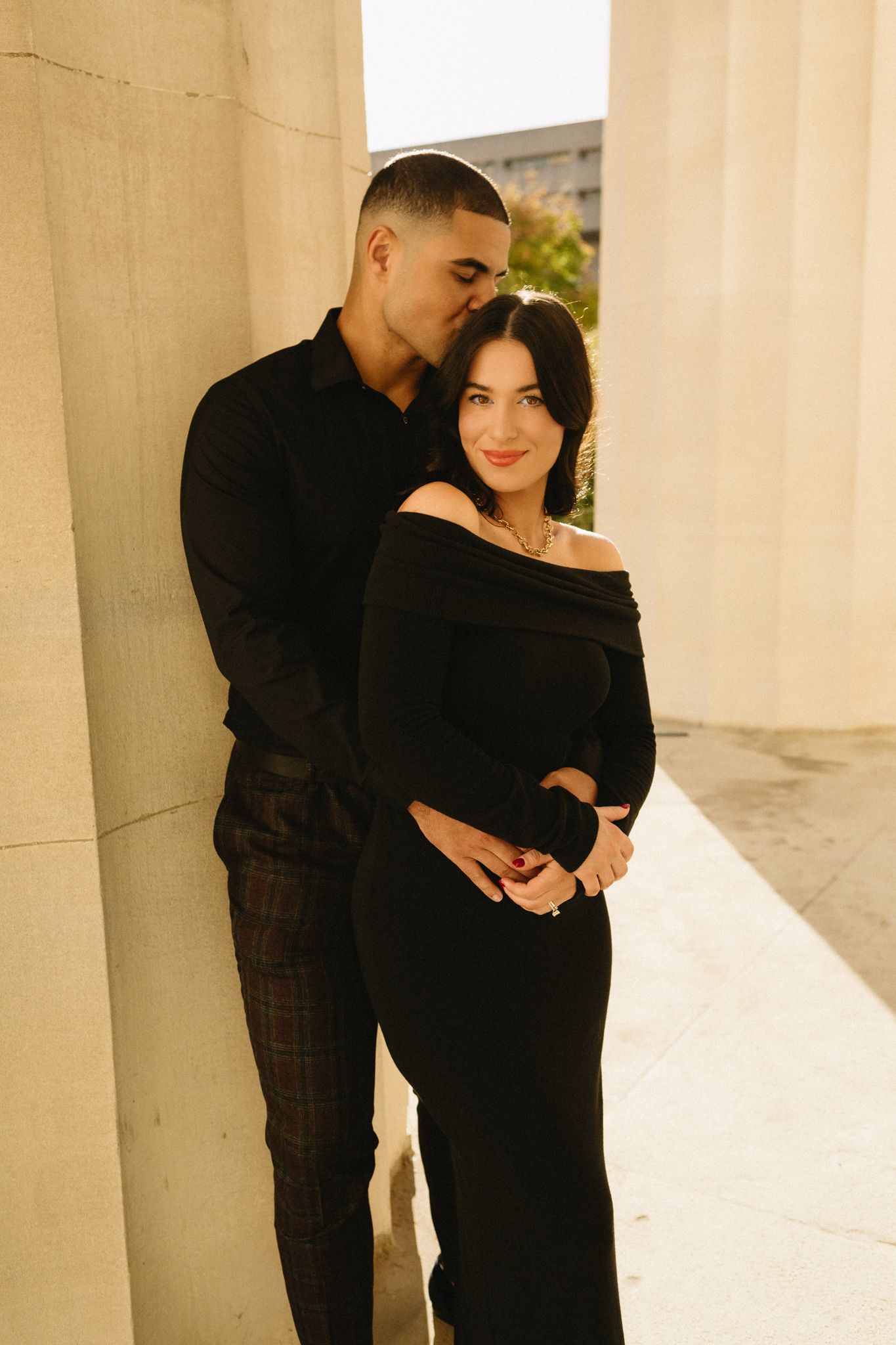 A stylish couple in matching black outfits pose together between elegant columns.