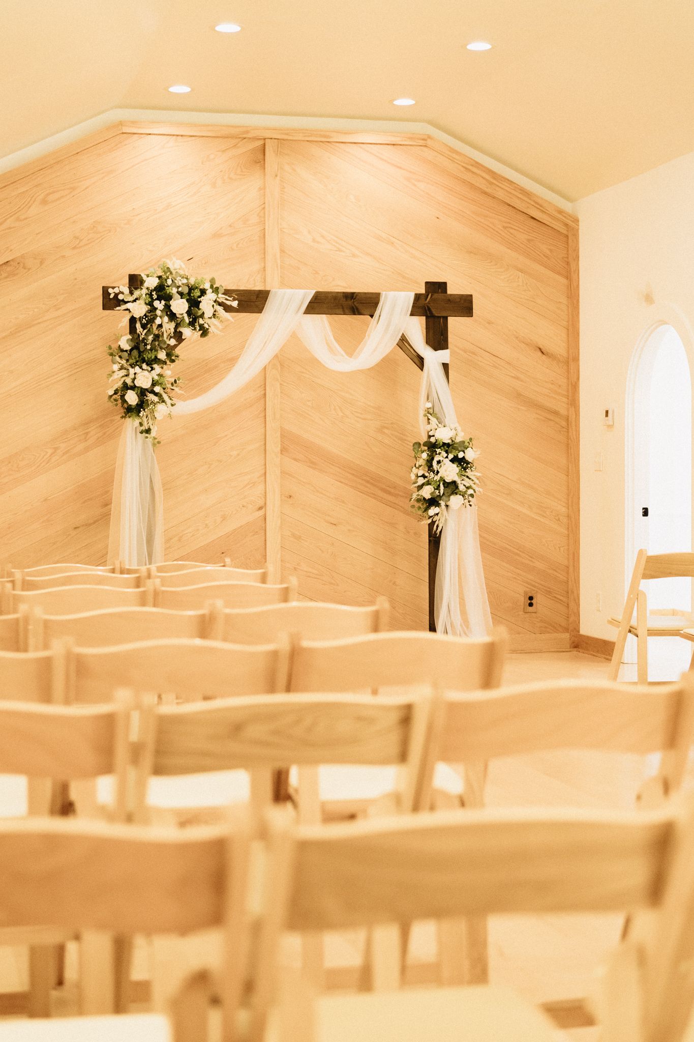Light wooden chapel interior with white drapery and floral arrangements adorning a cross altar at the end of rows of chairs.