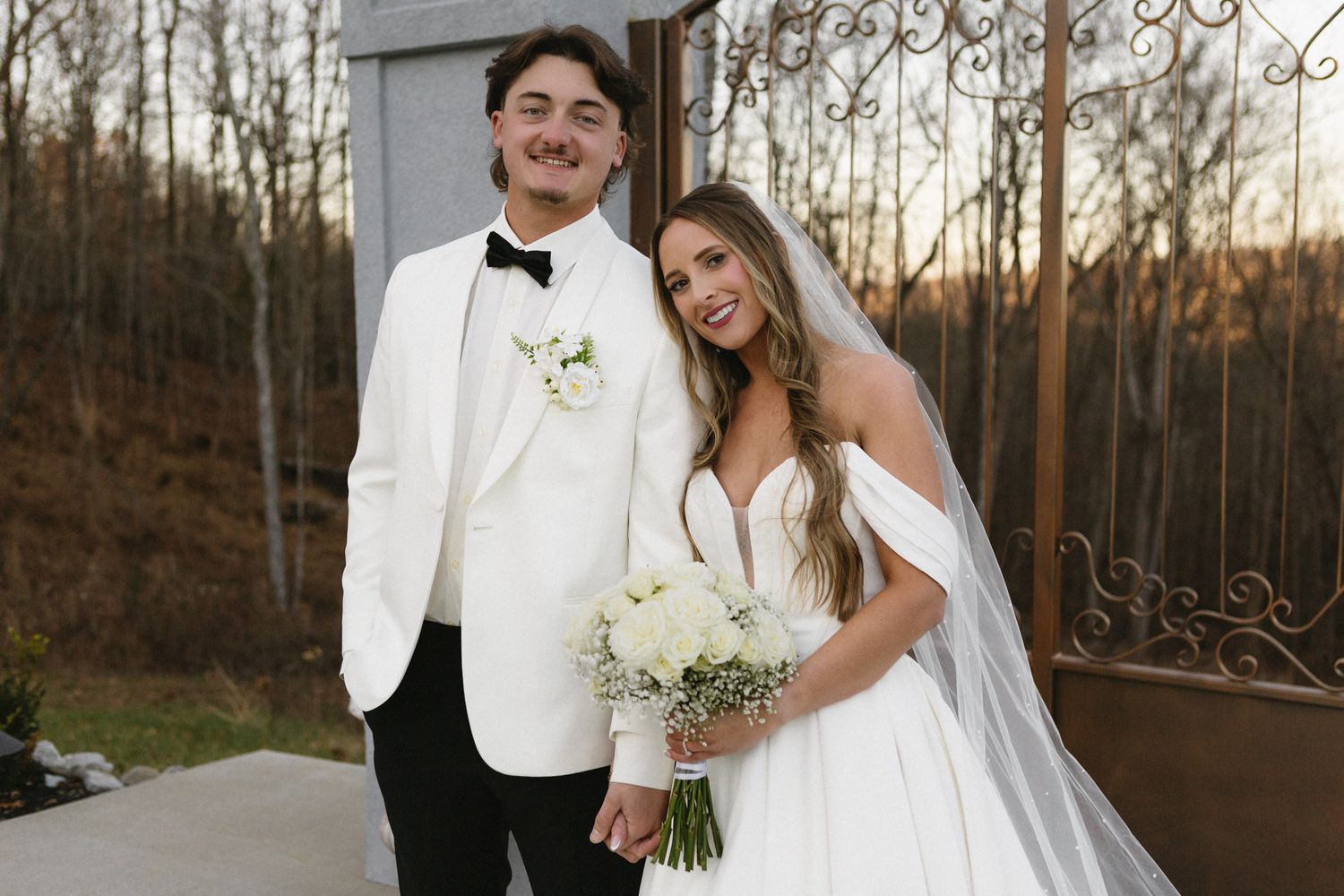Wedding couple in winter forest with bride in white gown and groom in white tuxedo holding flower bouquet.