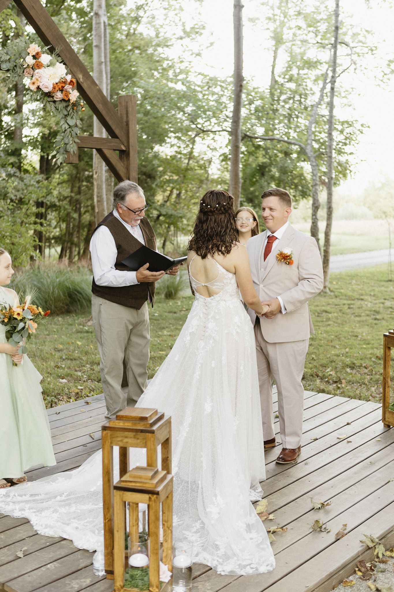 A rustic outdoor wedding ceremony takes place on a wooden deck surrounded by trees with lanterns and floral decorations.