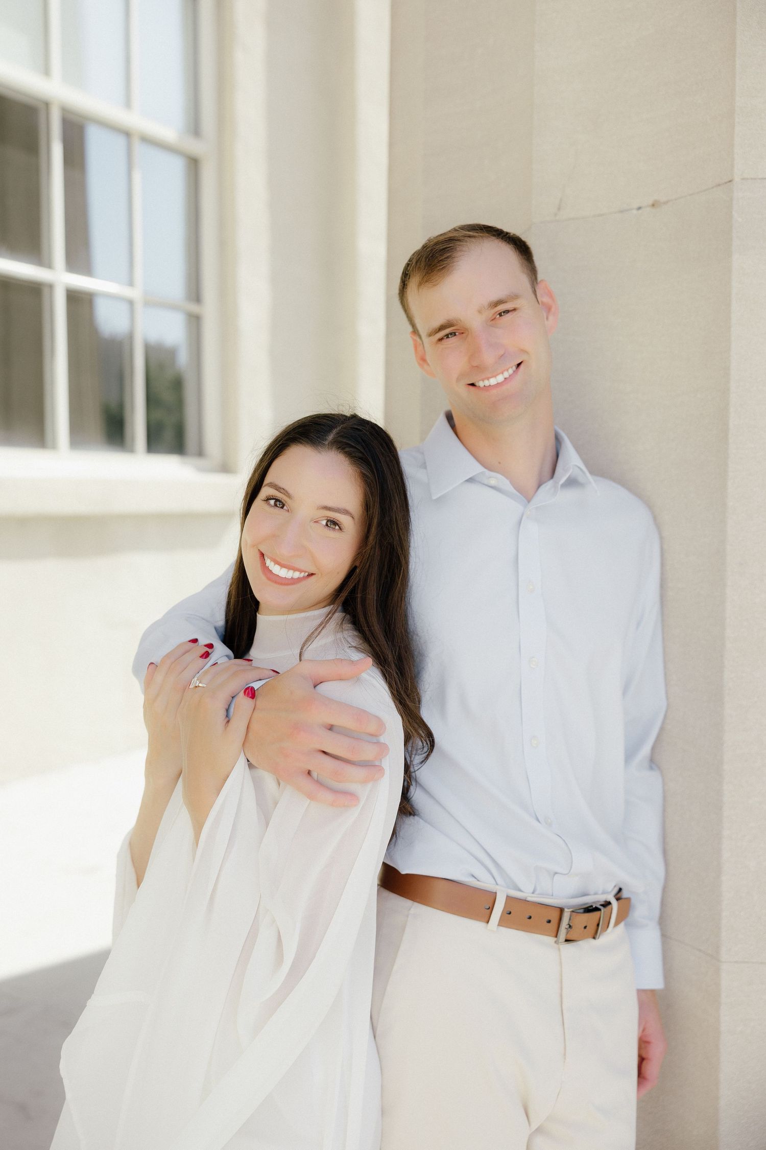 Smiling couple poses together against a white column, woman showing engagement ring in soft natural light.