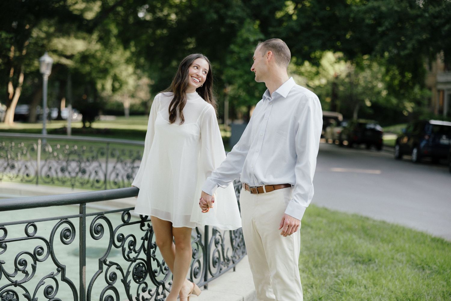 Happy couple holding hands and walking along an ornate iron fence in a lush green park.