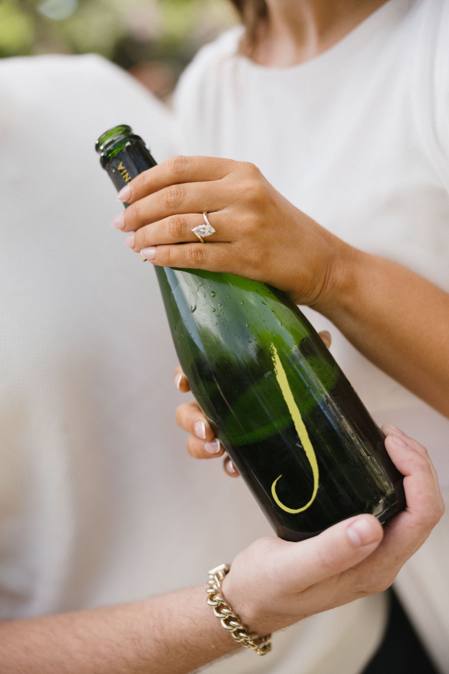 Two people holding a green champagne bottle monogrammed with letter J, one wearing an engagement ring.