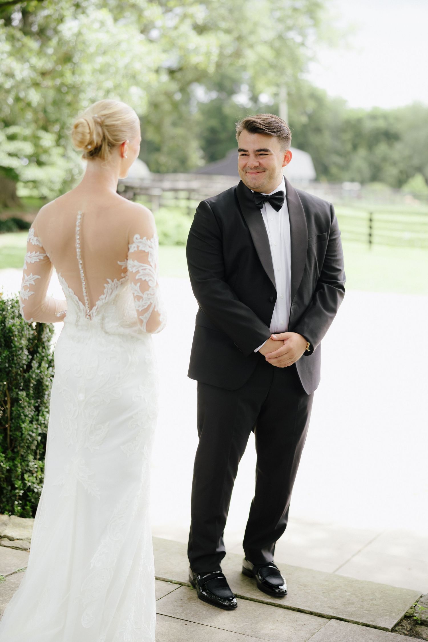Groom in black tuxedo smiling at bride in lace backless wedding dress during outdoor first look ceremony.
