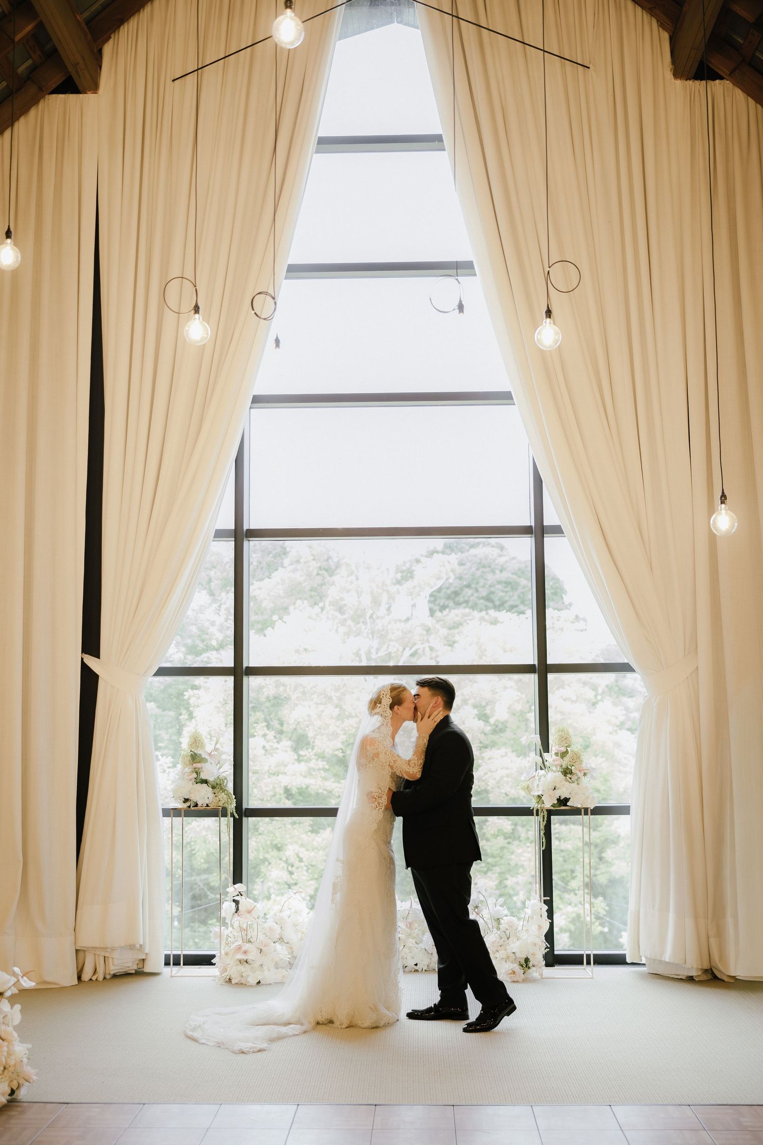 Bride and groom share a kiss at altar framed by ivory drapes and large window with garden views.