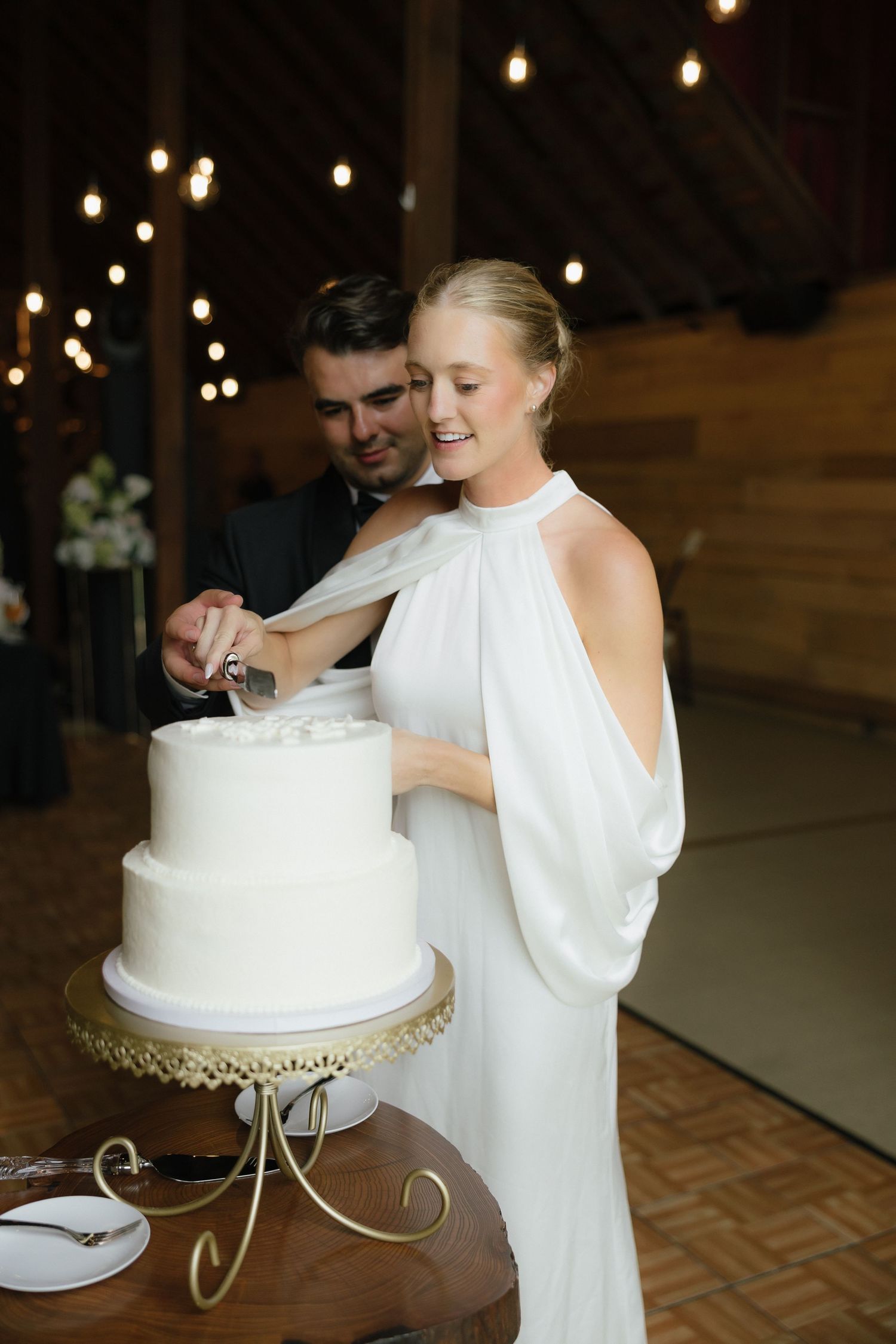 Smiling bride in elegant halter gown cuts two-tier white wedding cake with groom in warmly lit barn venue.