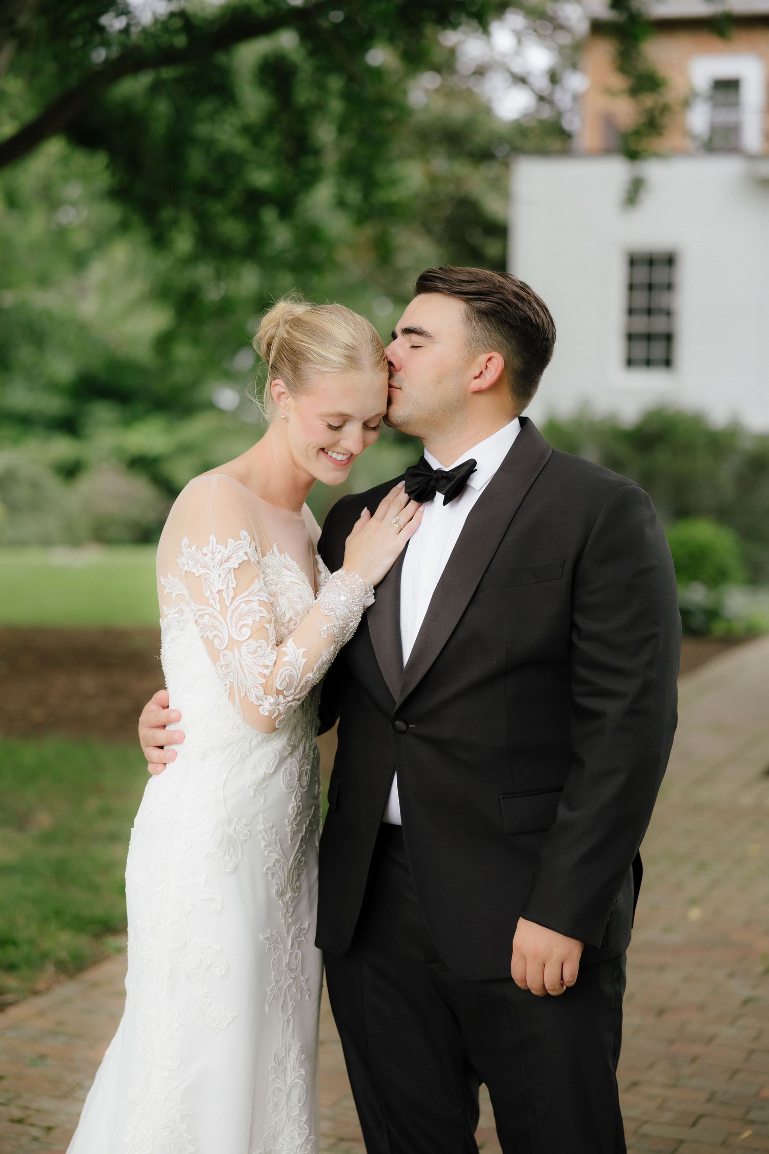 Groom in black tuxedo kisses bride in lace wedding dress on the cheek outdoors near a white building.