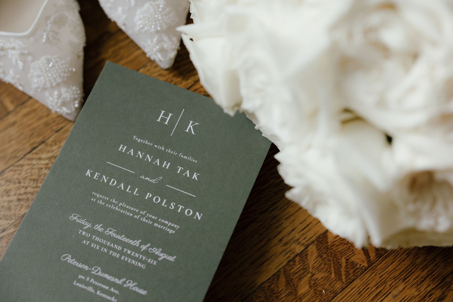 Elegant sage green wedding invitation for Hannah Tak and Kendall Polston with monogram, beside white floral bouquet.