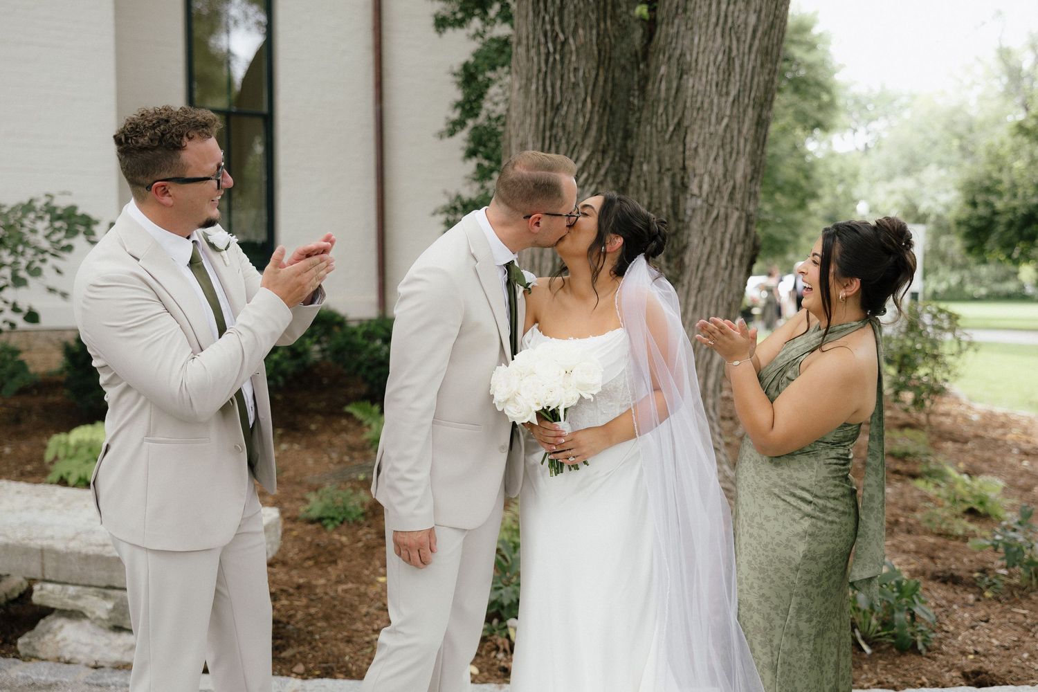 Bride and groom share a first kiss while guests celebrate beside a tree at outdoor wedding.