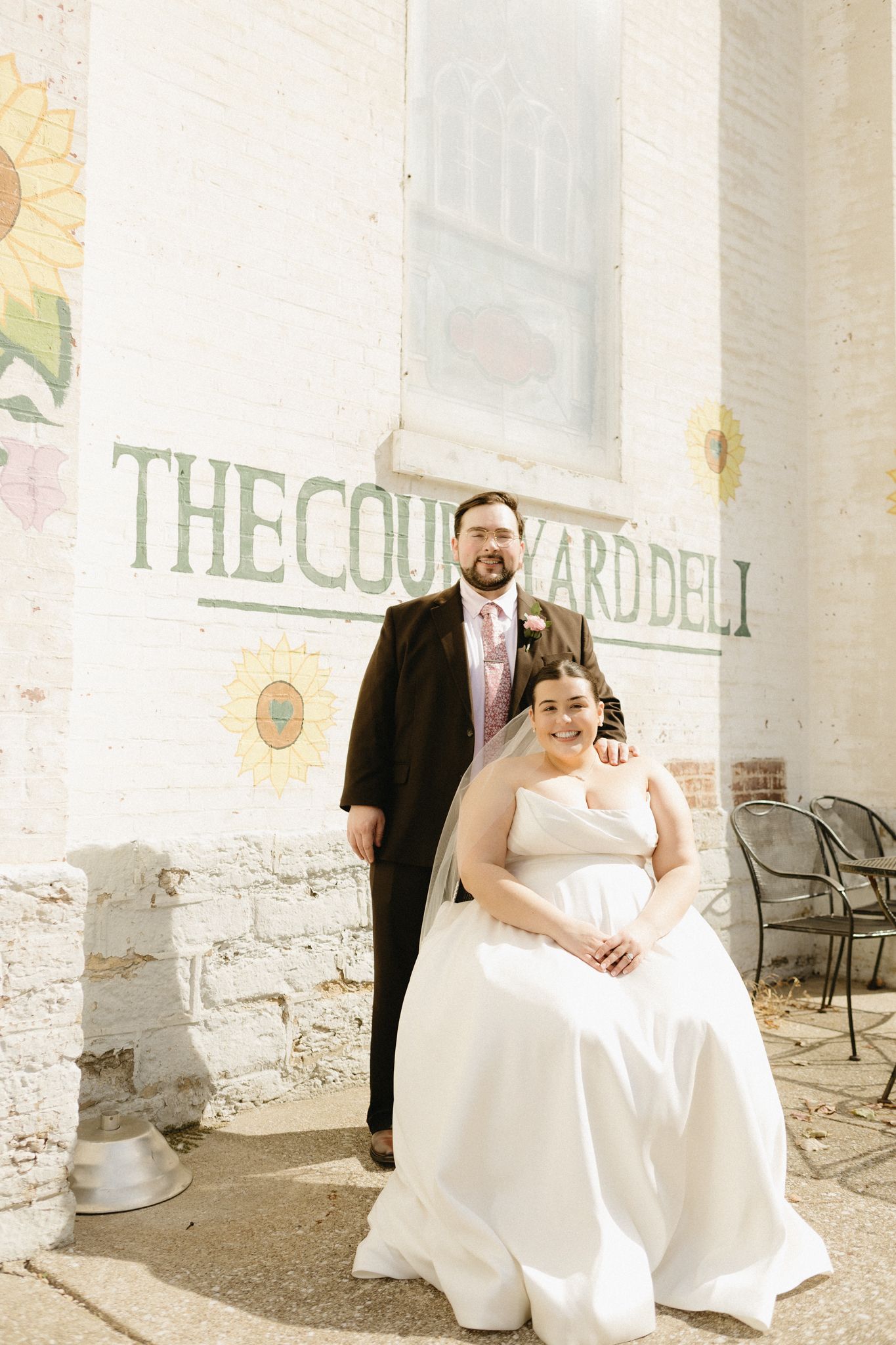 A wedding couple poses together in front of a rustic white wall with outdoor seating visible.