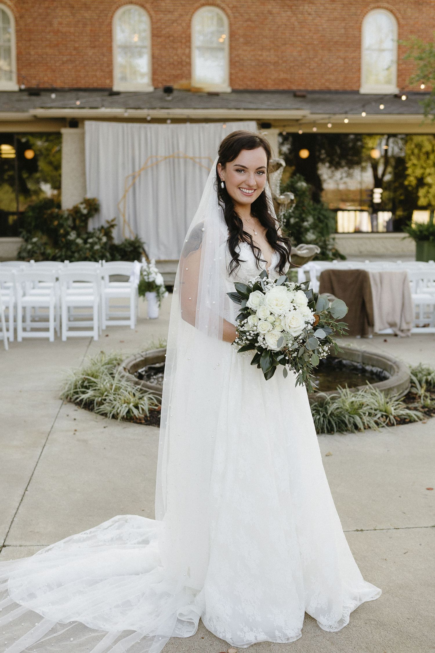 A bride in a classic white wedding gown with veil poses with her bouquet against a brick church backdrop.