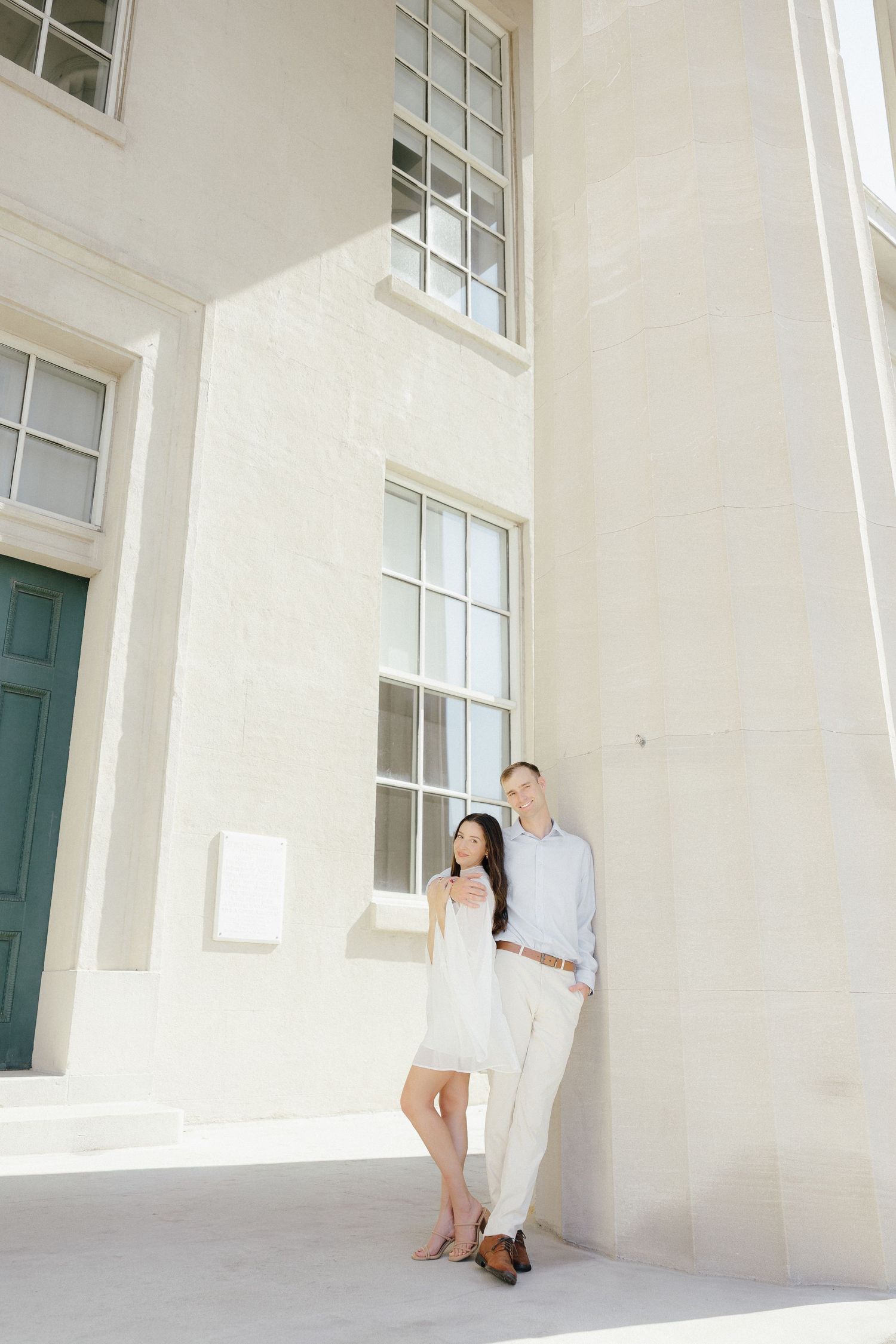Couple leaning together against a sunlit white stone building wall, woman in white dress smiling.