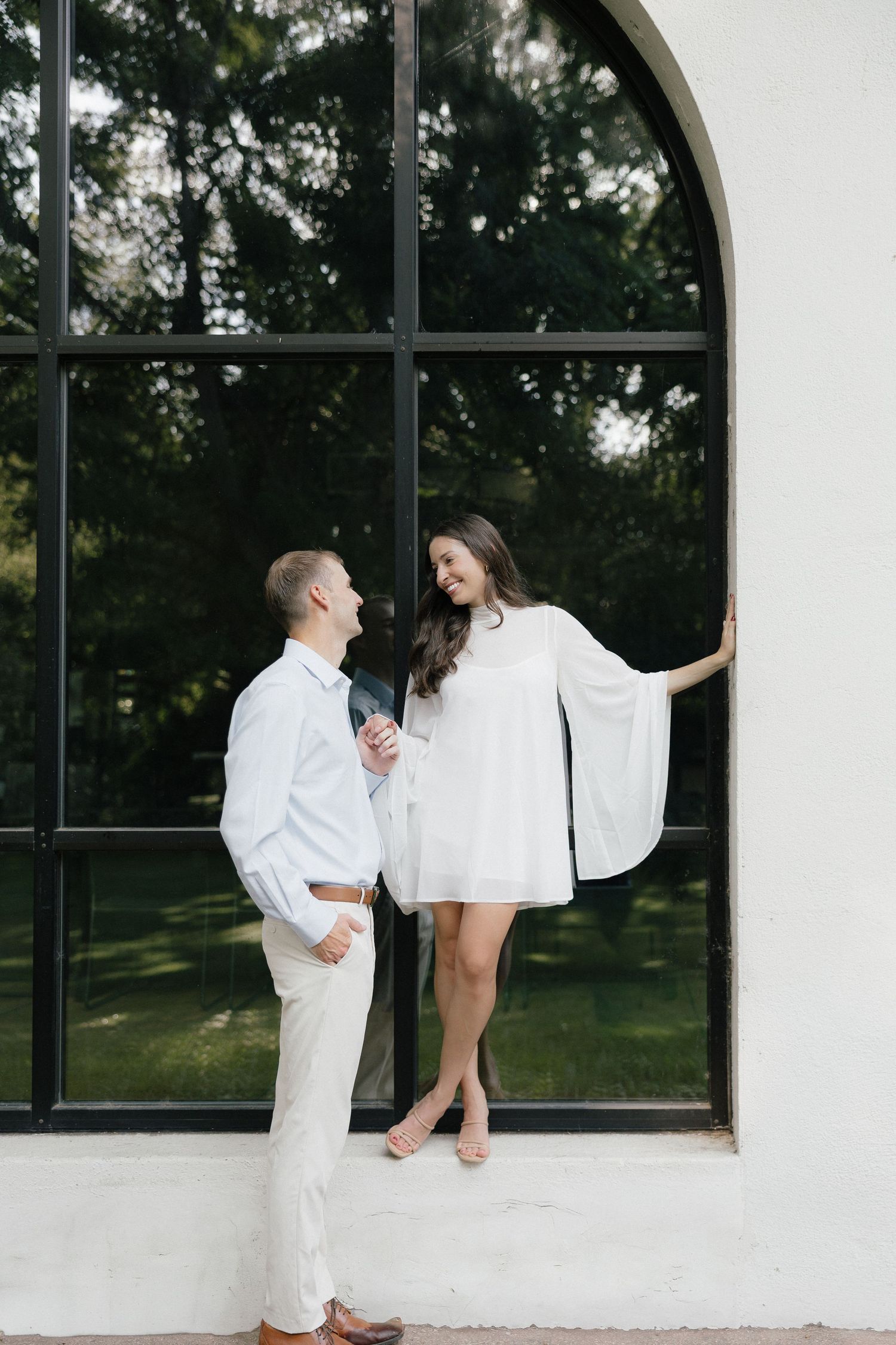 Couple smiling at each other by an arched window, woman in white cape dress leaning against white wall.