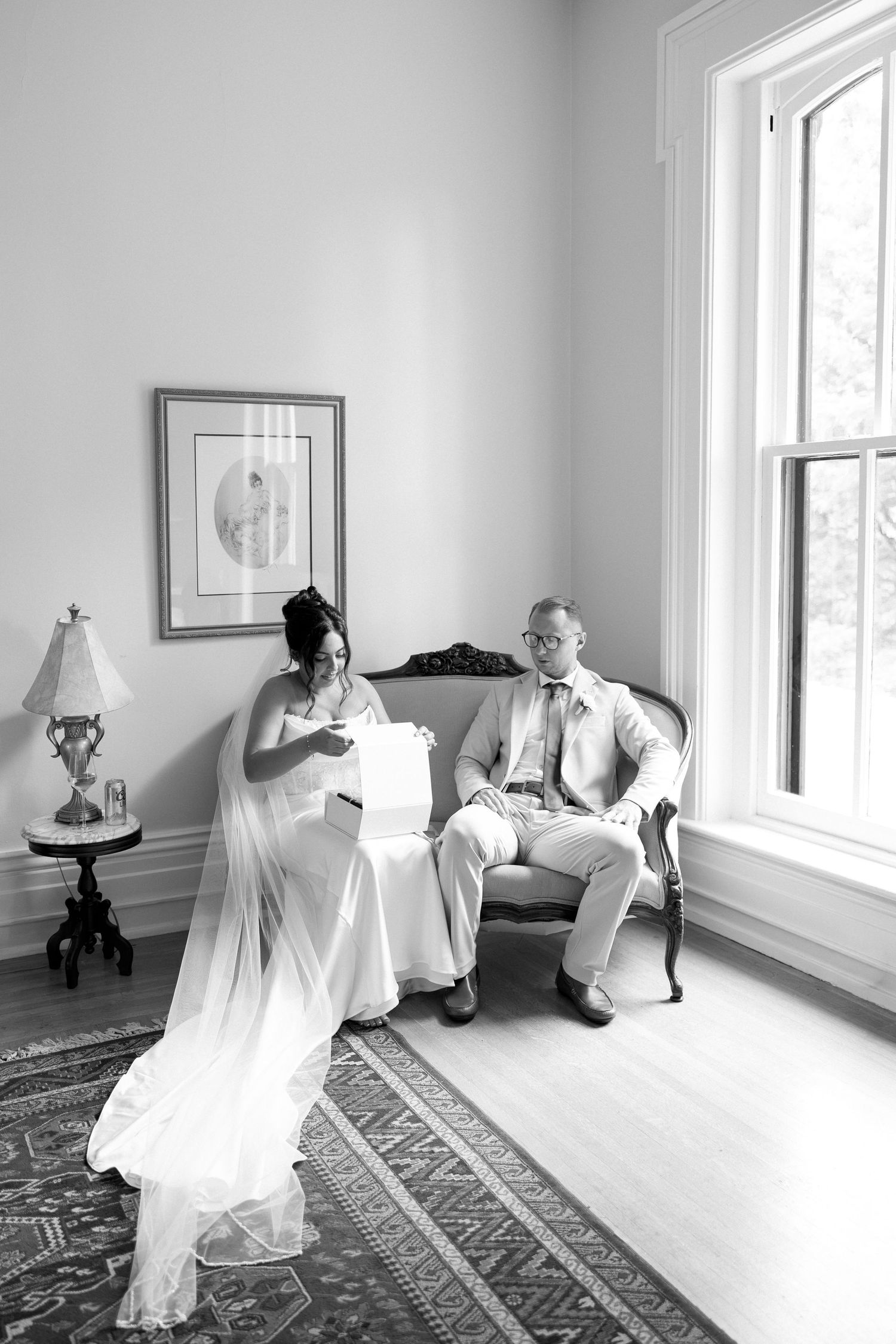 Black and white photo of bride and groom exchanging gifts while seated on vintage sofa indoors.