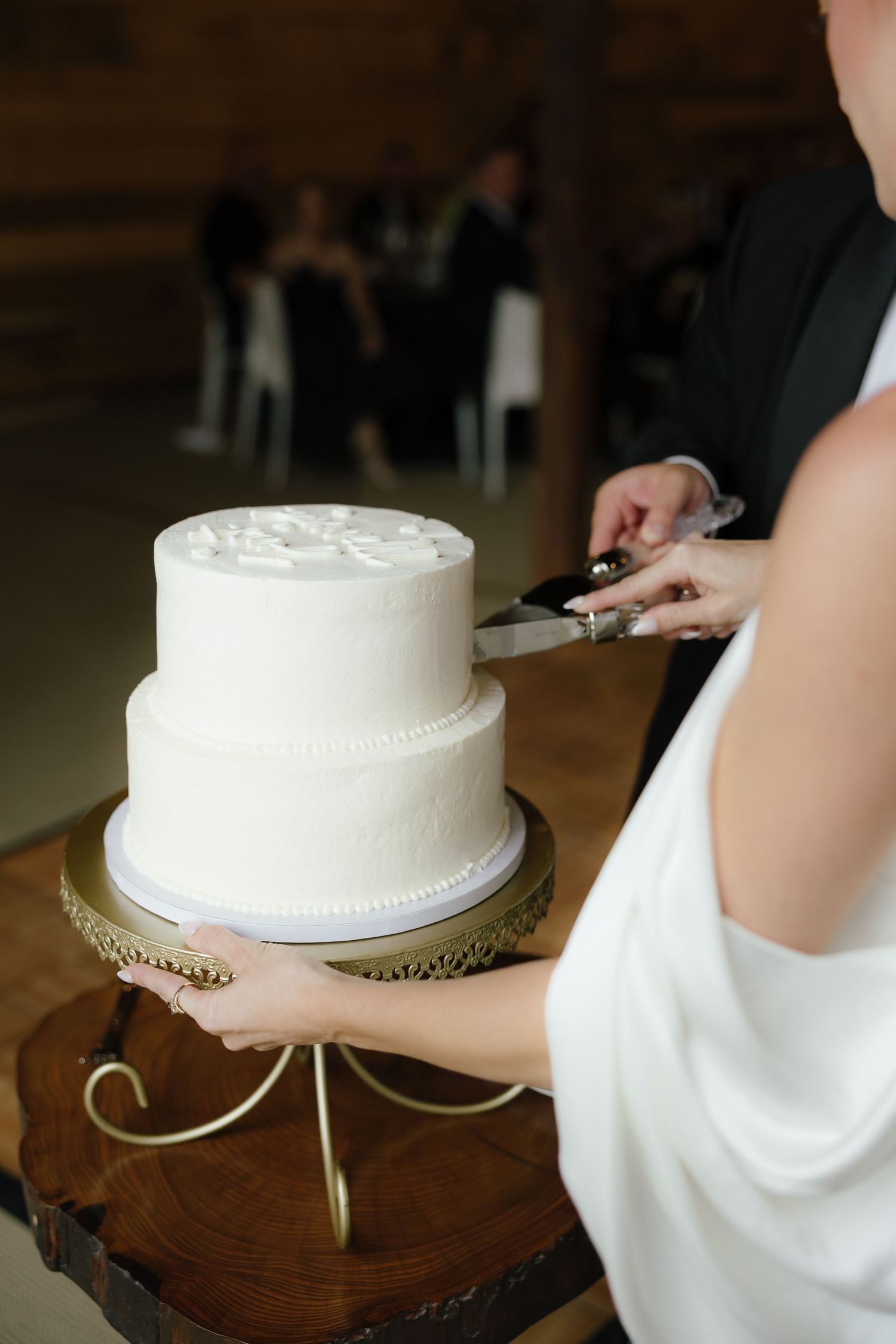 Bride and groom cutting elegant two-tier white wedding cake on gold stand at rustic barn reception.