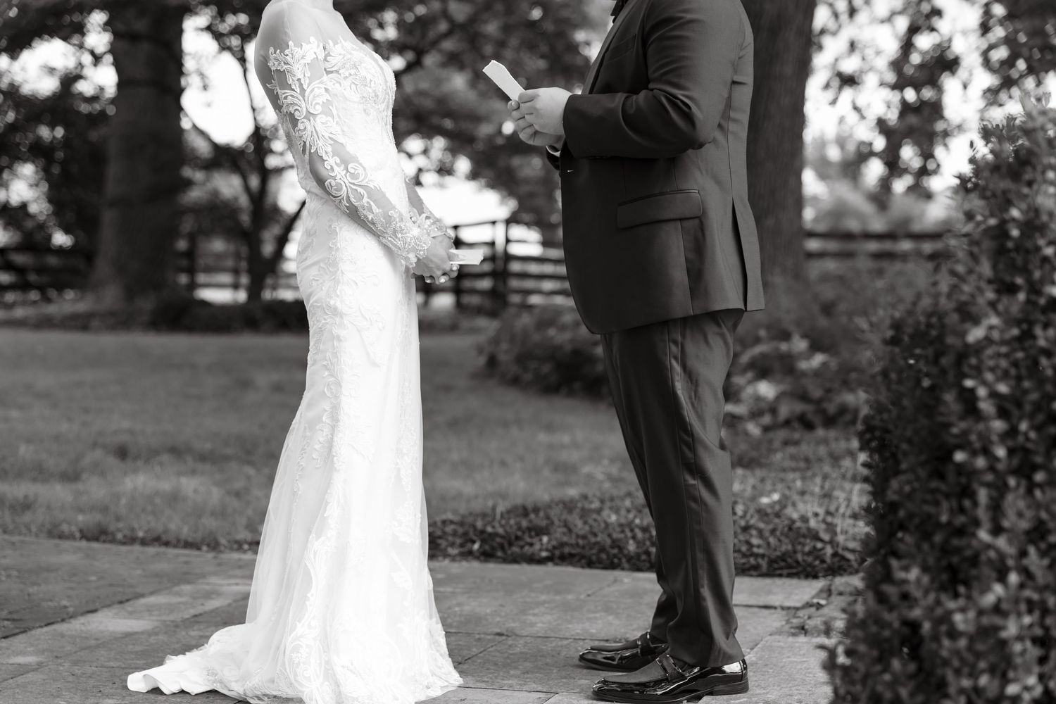 Black and white photo of bride in lace gown and groom in suit exchanging vows outdoors.